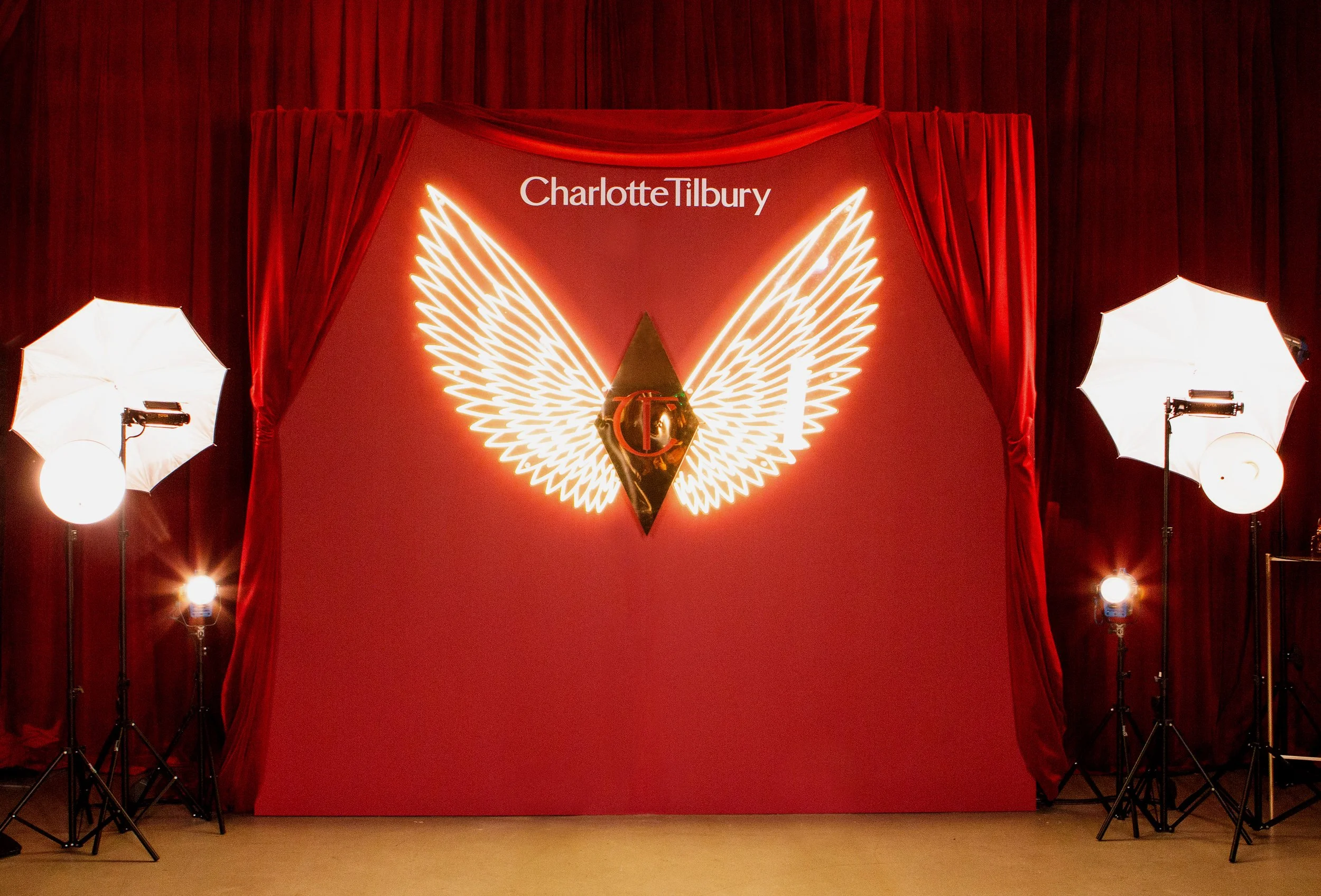 Charlotte Tilbury branded photo backdrop with illuminated neon angel wings on a red velvet stage