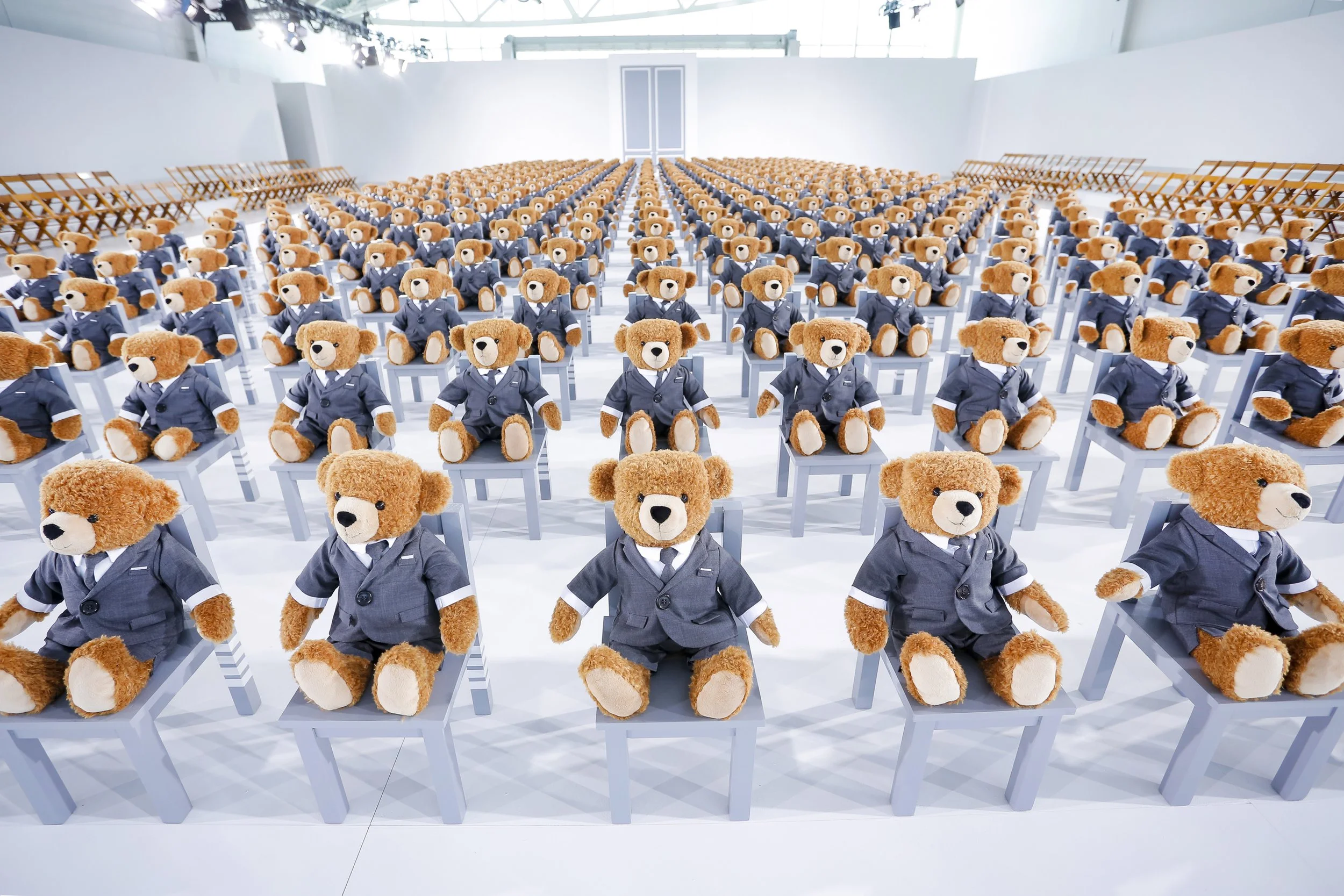 Hundreds of Thom Browne teddy bears in grey suits seated in rows of chairs at a fashion show venue