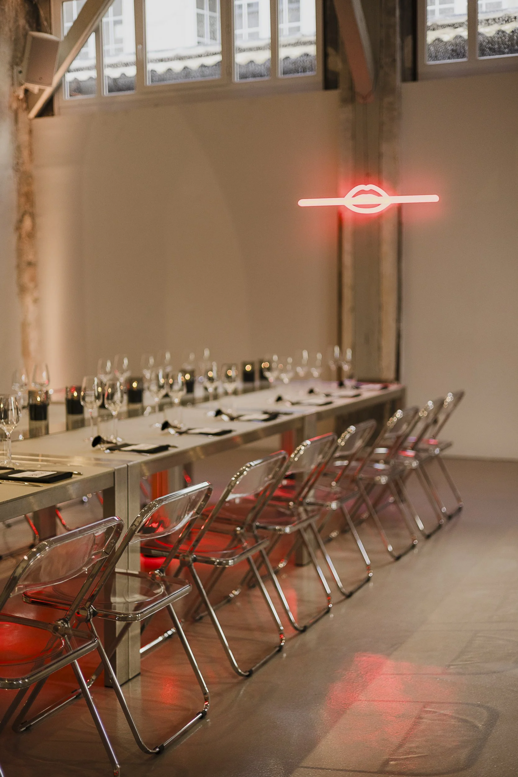 Long dinner table with acrylic ghost chairs and a neon red lips sign in an industrial loft space