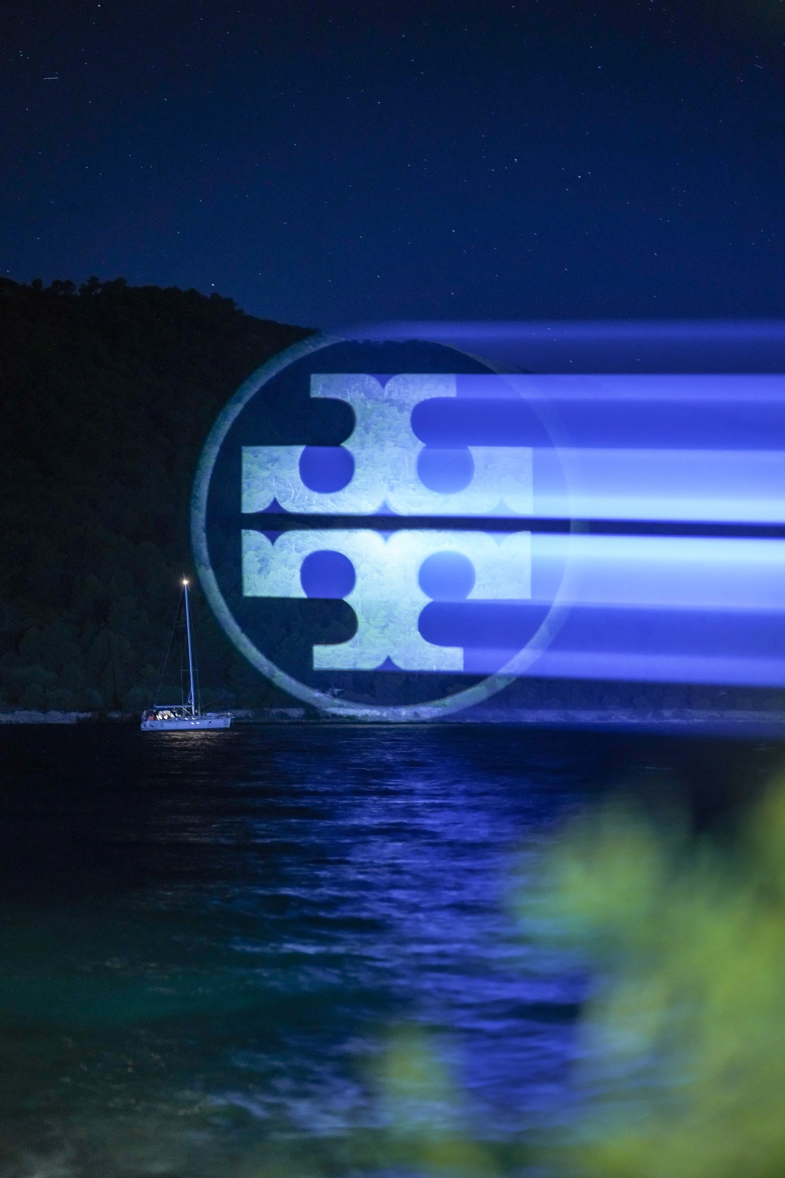 Tory Burch logo projected as a blue light beam over dark sea at night with a sailboat behind
