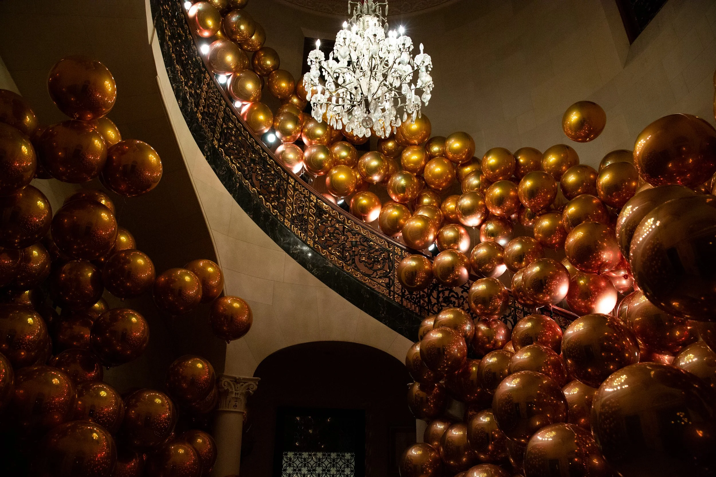 Ornate curved staircase filled with gold chrome balloon clusters beneath a chandelier