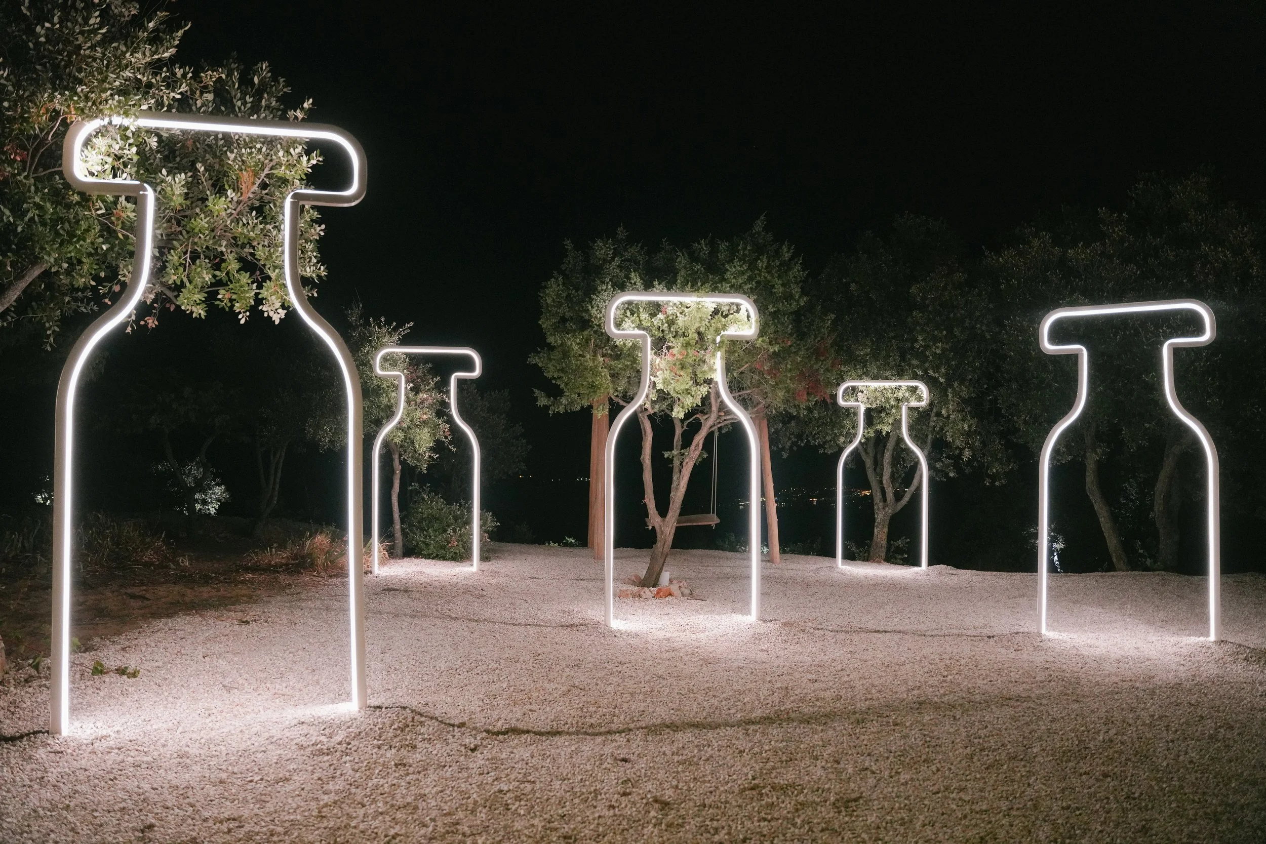 Neon perfume-bottle outlines lit at night among olive trees
