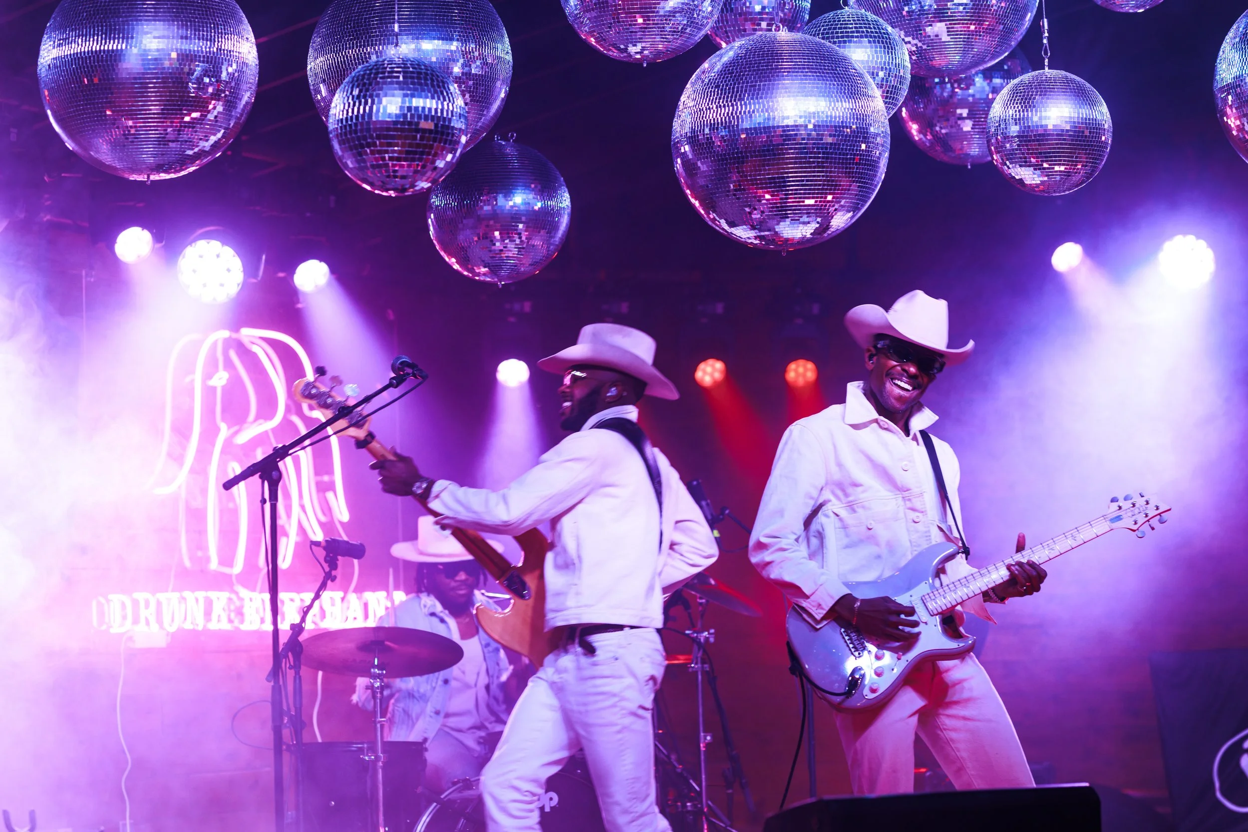 Live band in white cowboy outfits performing on stage under mirror disco balls at a purple-lit event