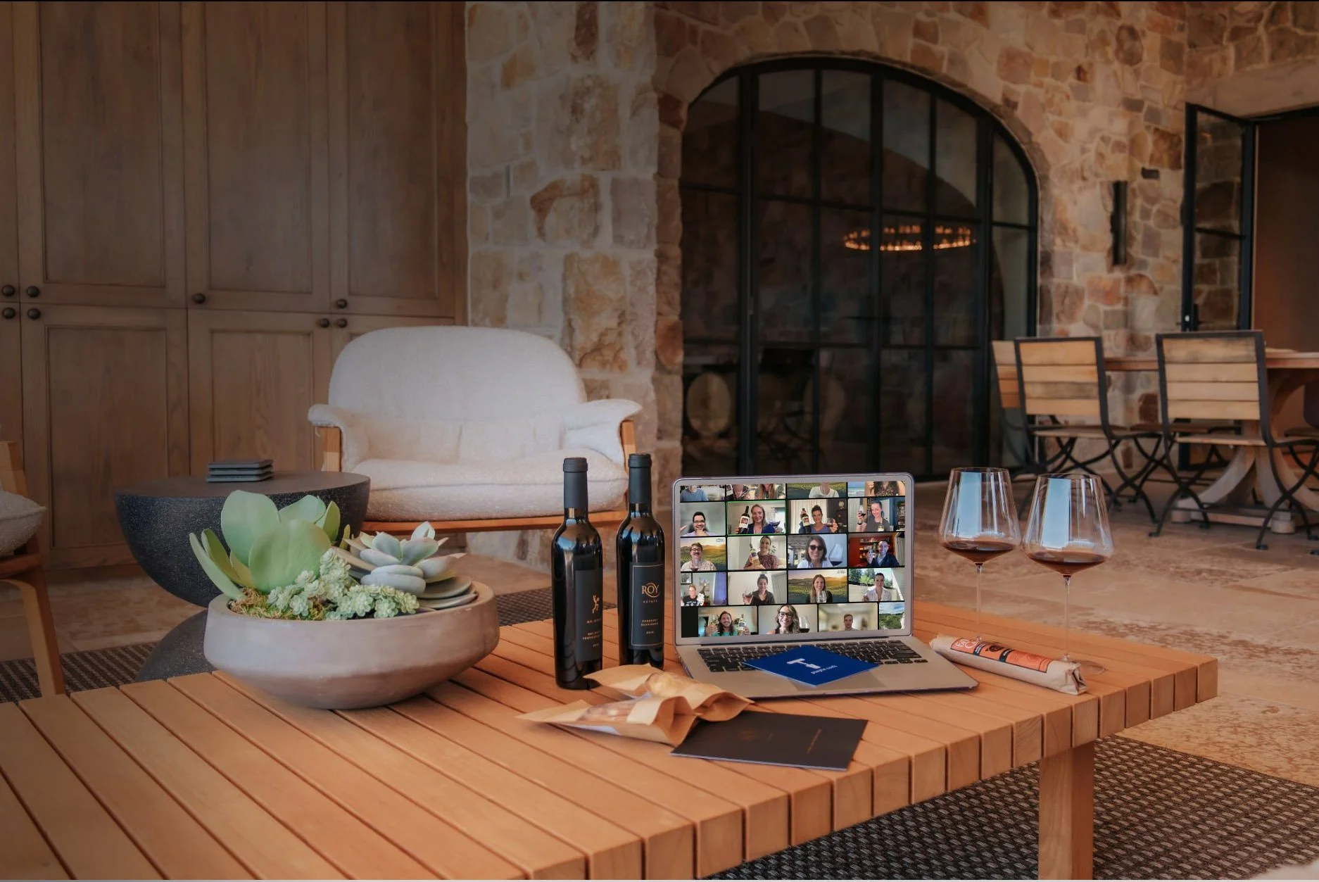 Wine bottles beside a laptop showing a live virtual tasting on a rustic winery table