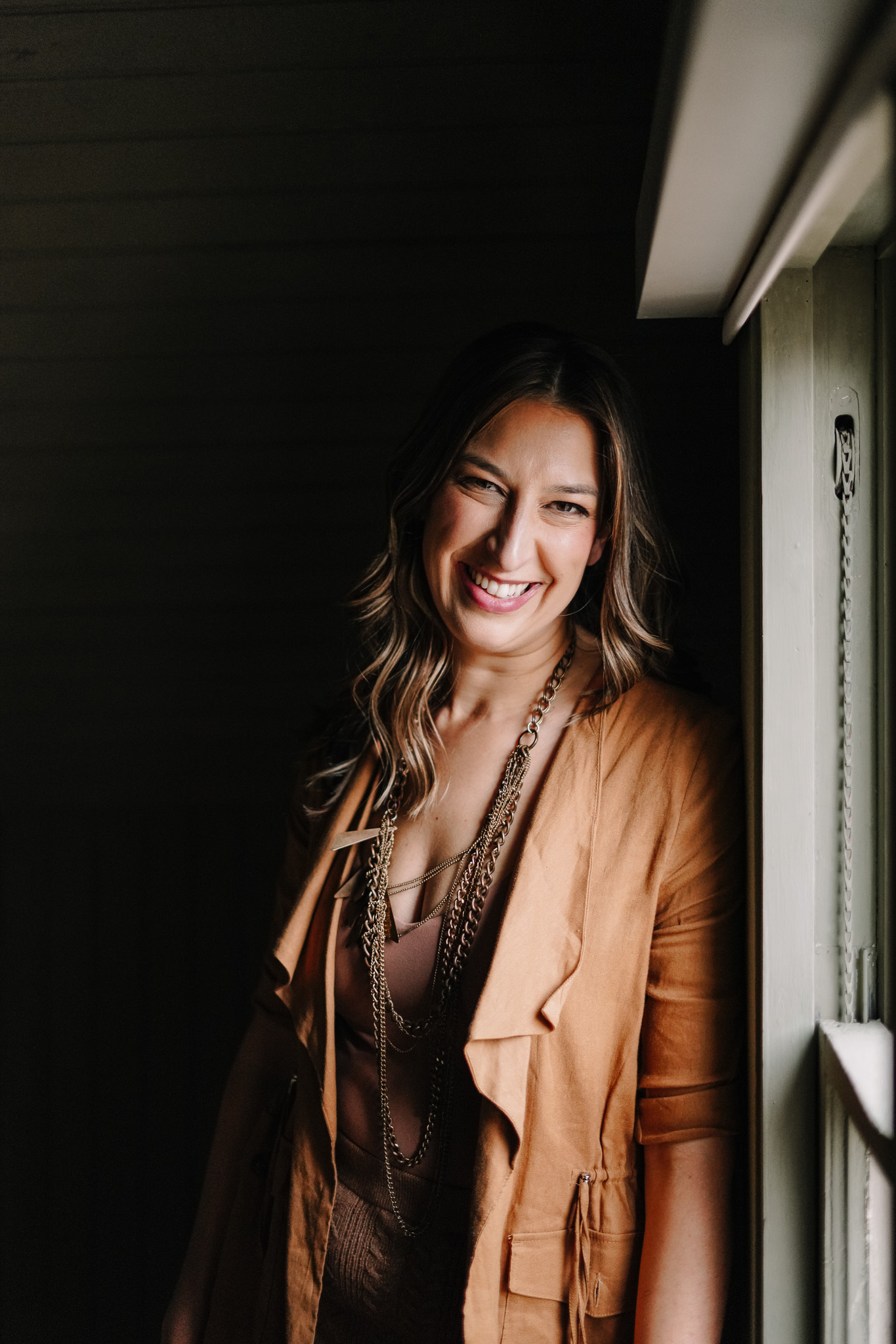 Emily Fowler Pellen, smiling in a tan caramel jacket, leaning against a window frame