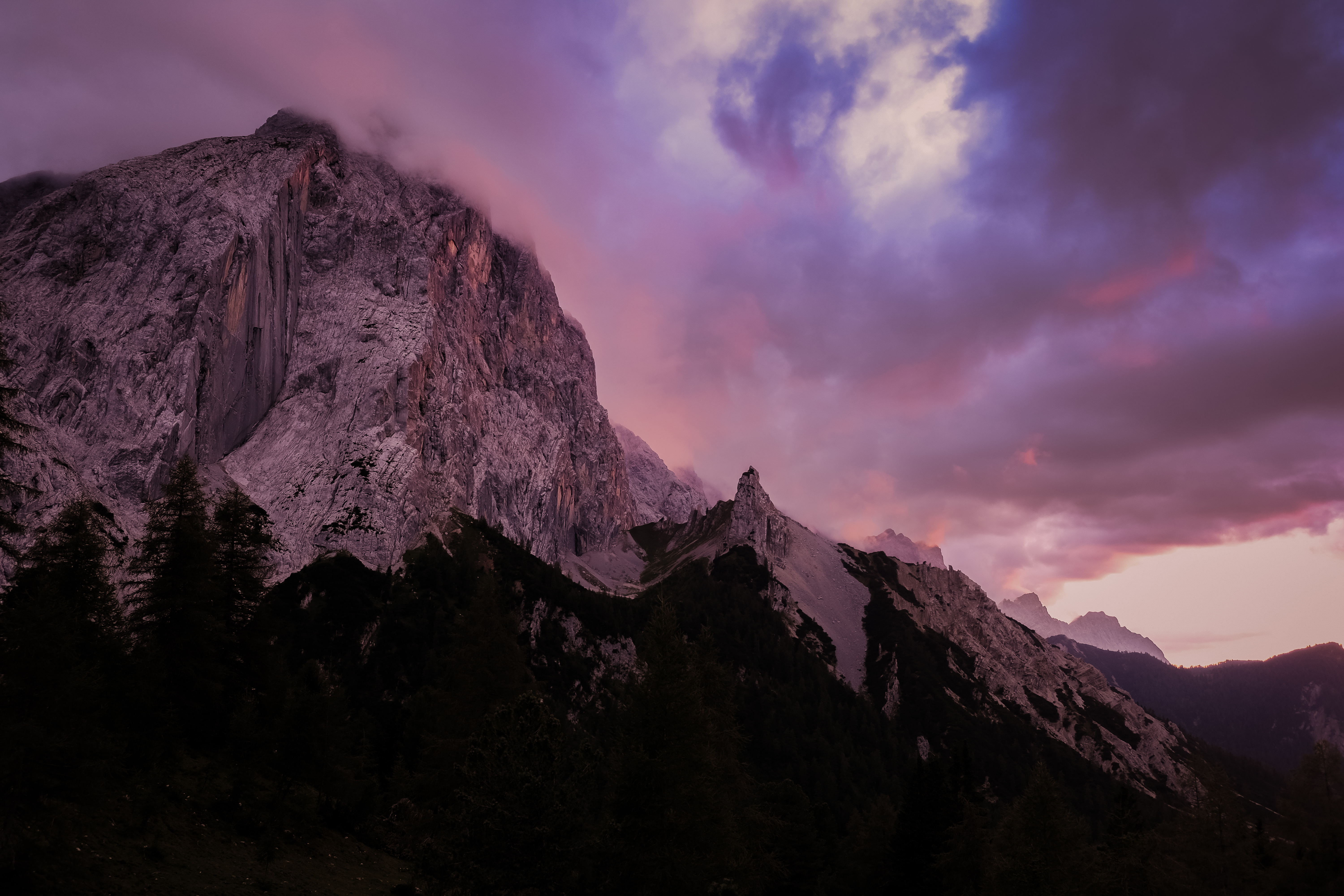 Dramatic mountain peak beneath a purple and pink sunset sky, dark treeline silhouette in the foreground