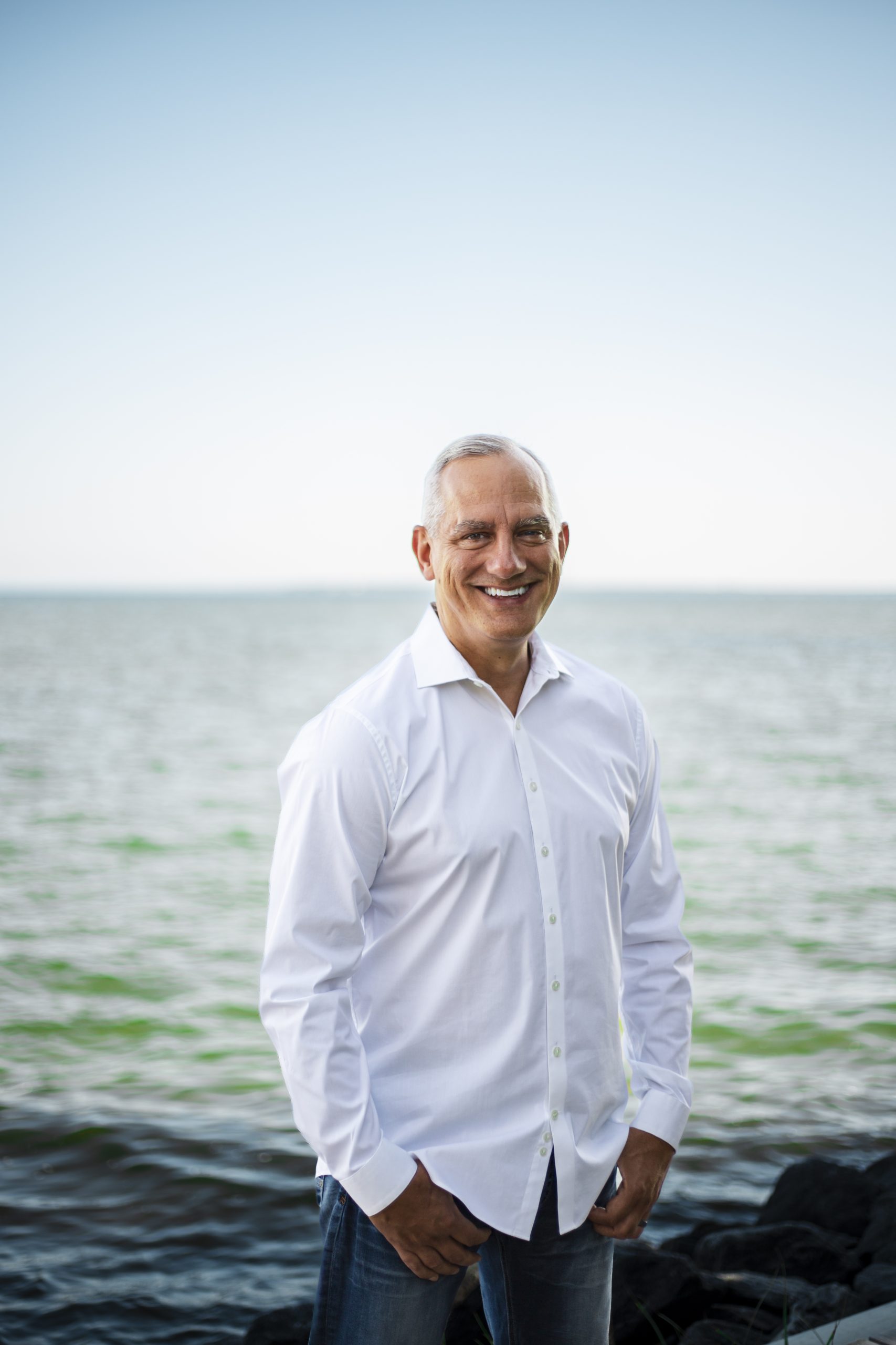 Joe Mikalajunas in a white button-up shirt standing on a rocky shoreline with the ocean behind him