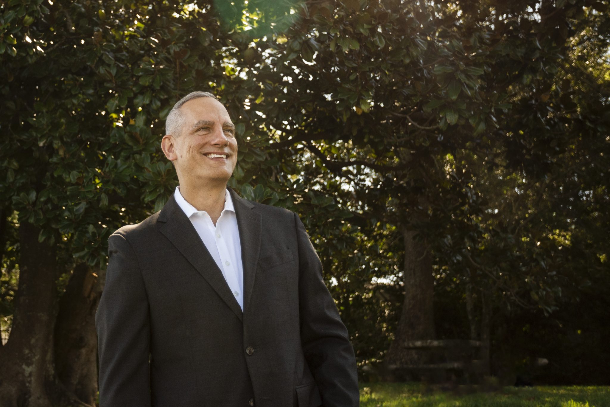 Joe Mikalajunas in a dark suit jacket looking upward and smiling beneath sunlit trees