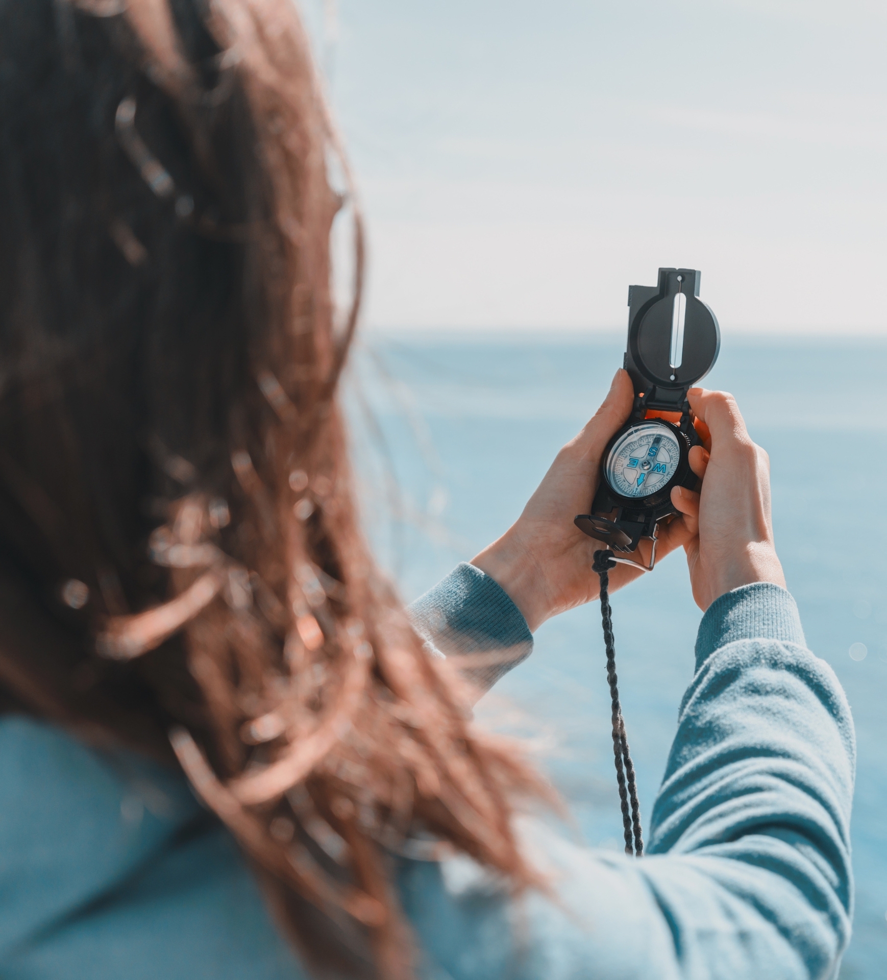 A person seen from behind holding a military-style compass toward the ocean horizon