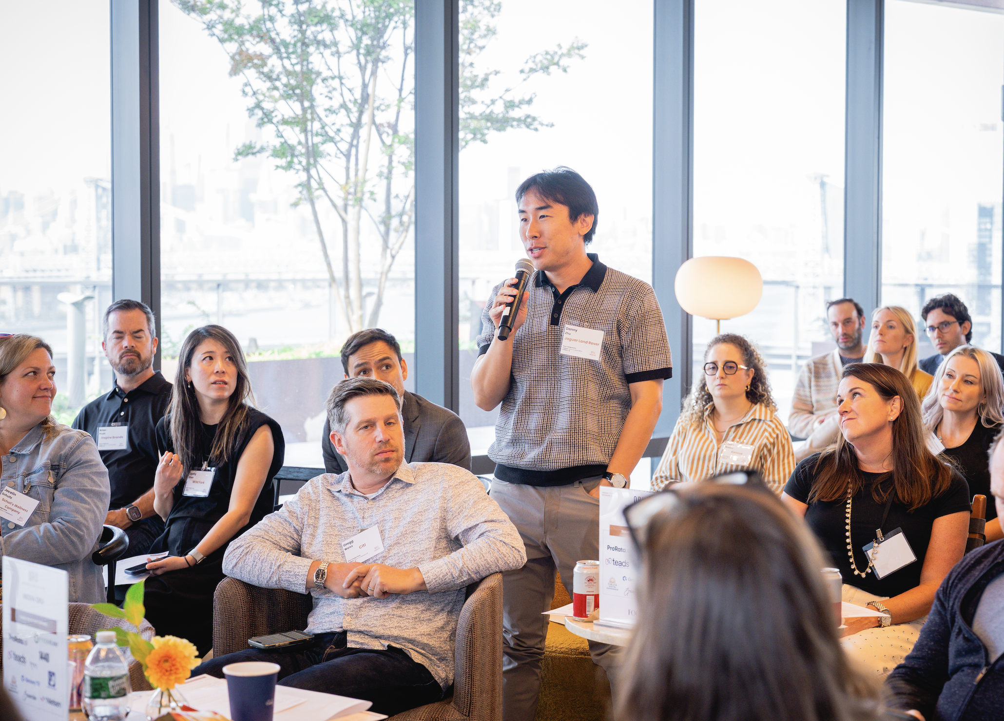 Attendee standing and speaking into a microphone amid a seated group of executives