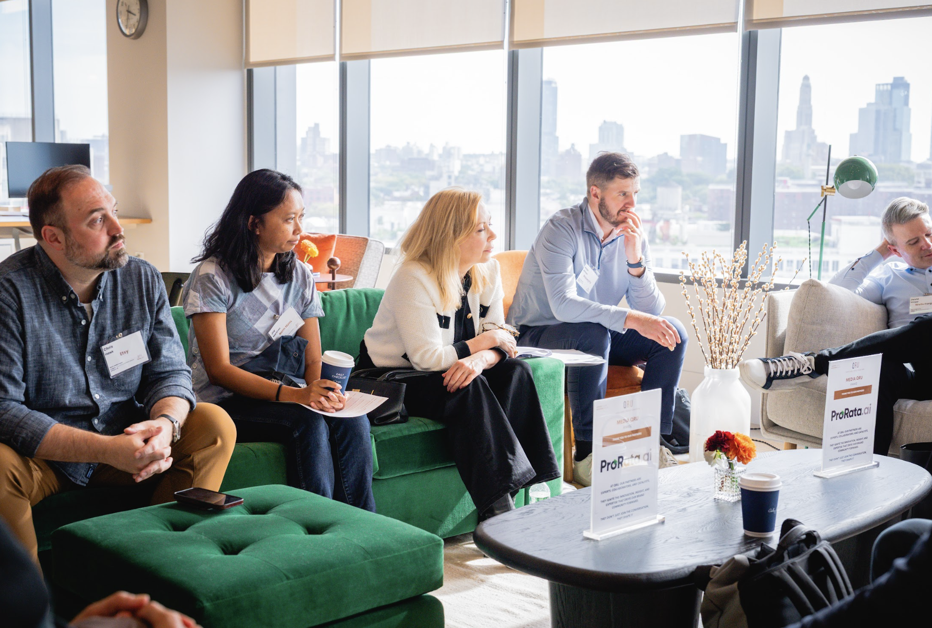 Five attendees seated on green sofas listening, ProRata.ai table cards, city skyline behind
