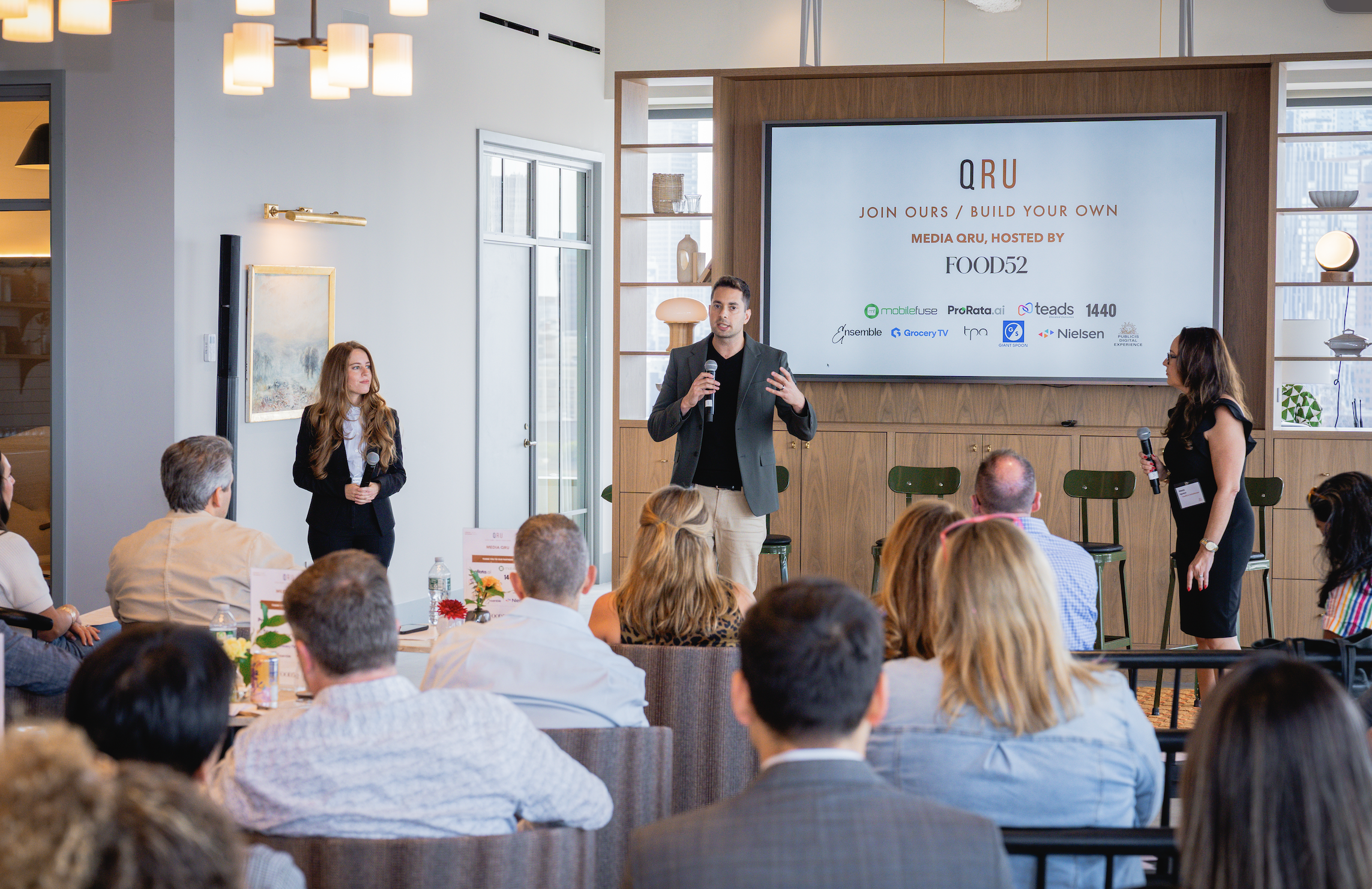 Three presenters standing at the front of the room with a QRU branded slide, seated audience at tables