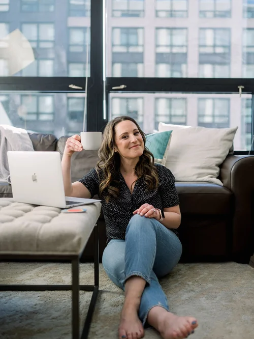 Rachel Naar seated on the floor of a sunlit apartment beside a laptop, holding a coffee mug, with city windows behind her