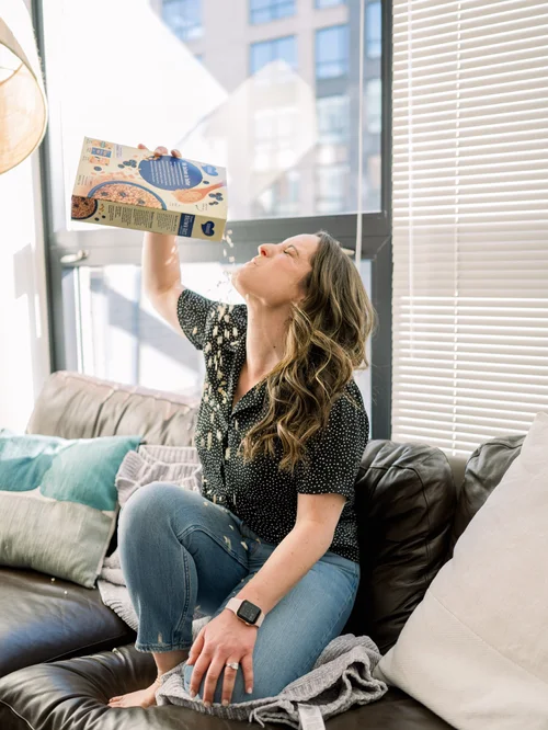 Rachel Naar on a sofa holding a food package up to the window light, reading the label