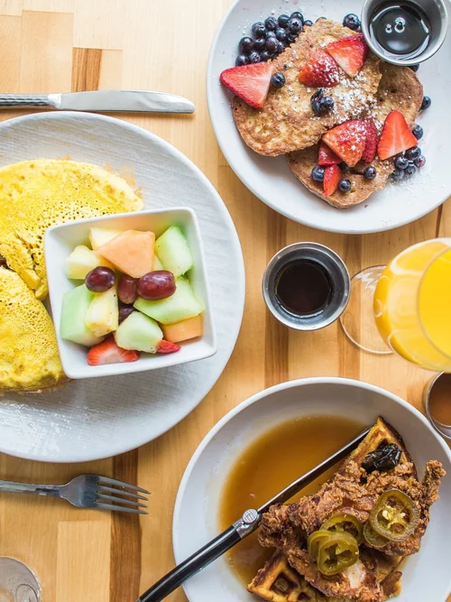 Overhead brunch spread with pancakes and berries, eggs, fruit, chicken and waffles, and orange juice