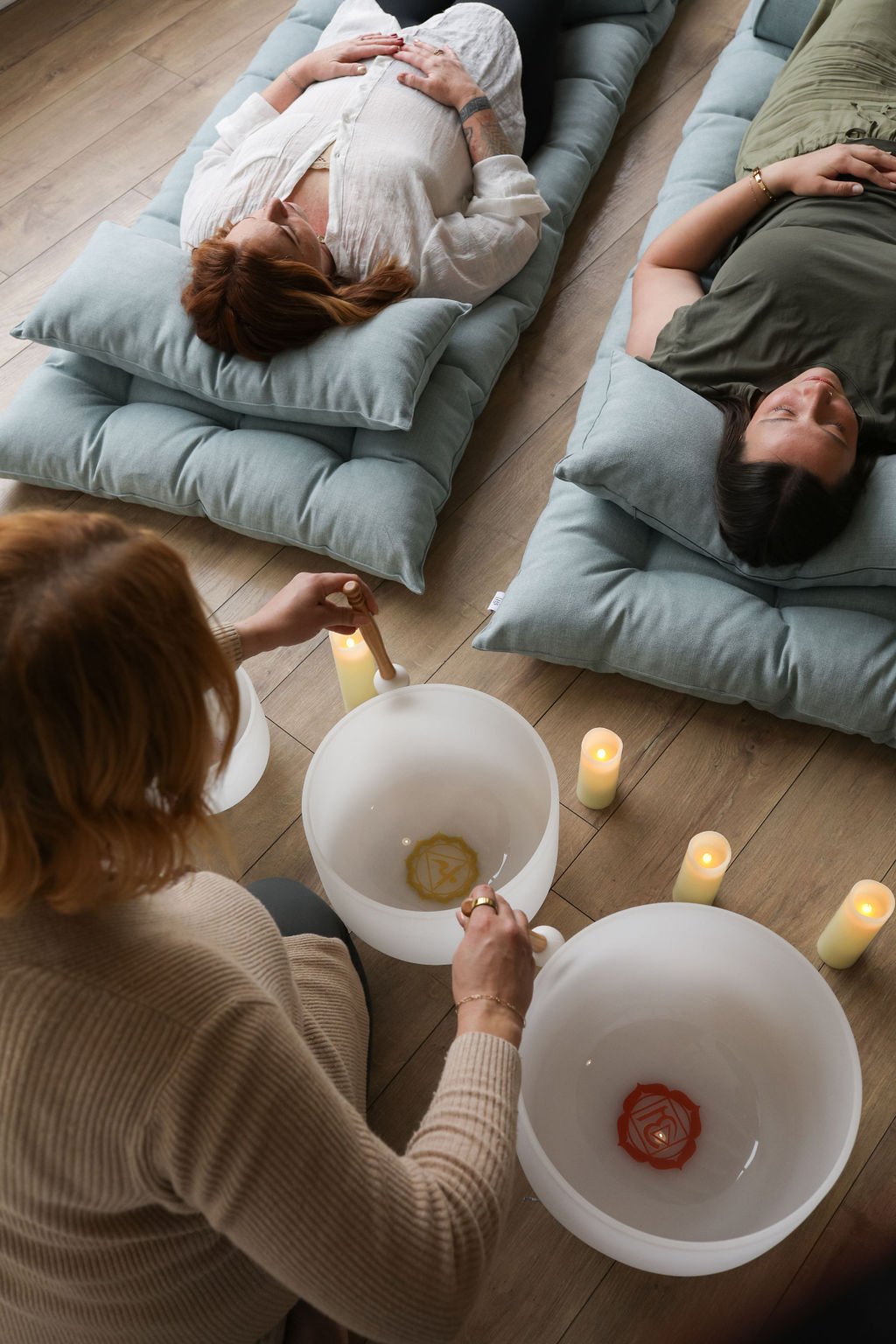 Overhead view of a sound bath session: a practitioner playing crystal singing bowls with candles while two participants rest on teal floor cushions