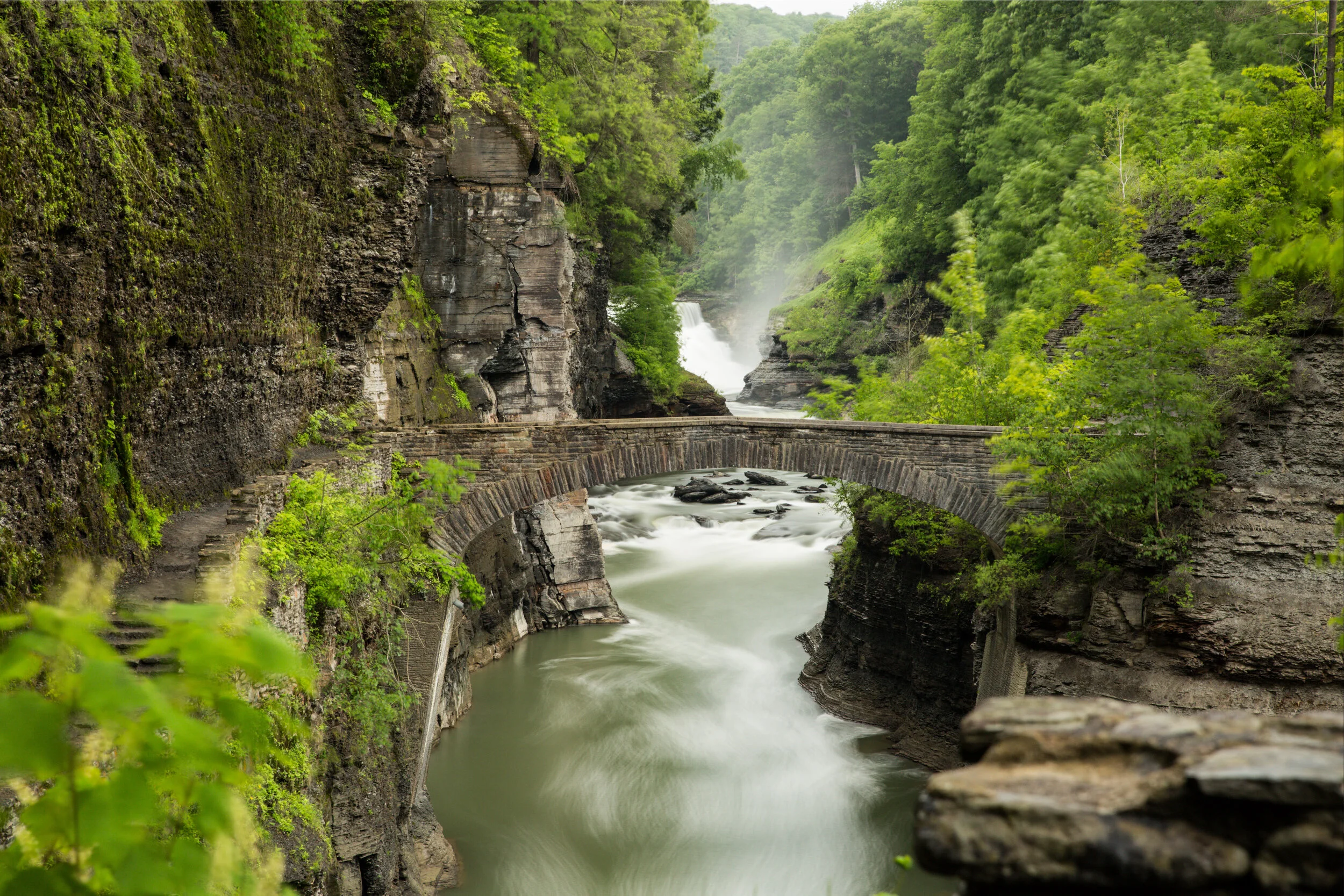 Stone arch bridge over a rushing river in a lush green gorge with a waterfall behind