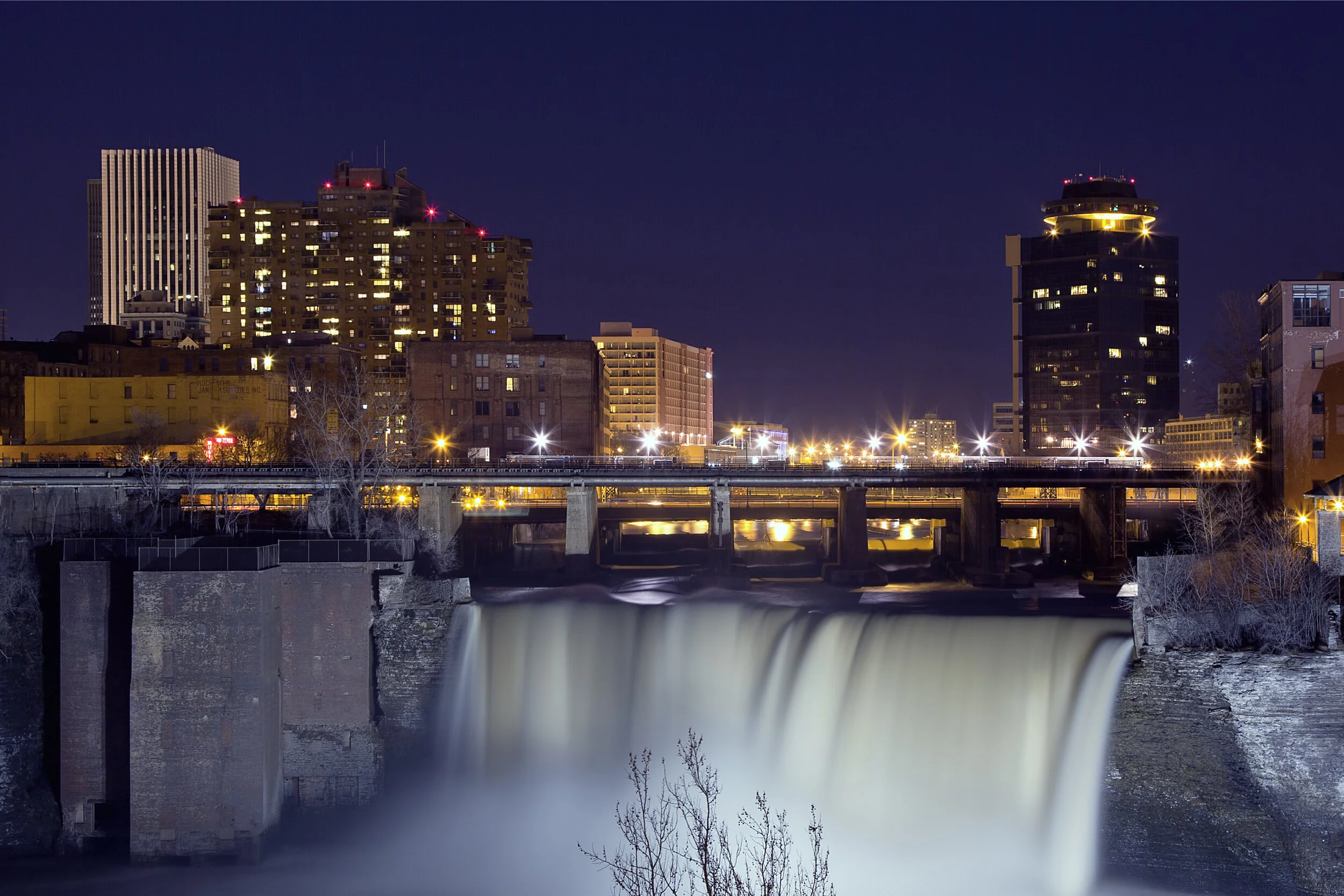 Rochester skyline at night with High Falls waterfall lit in the foreground and an illuminated bridge