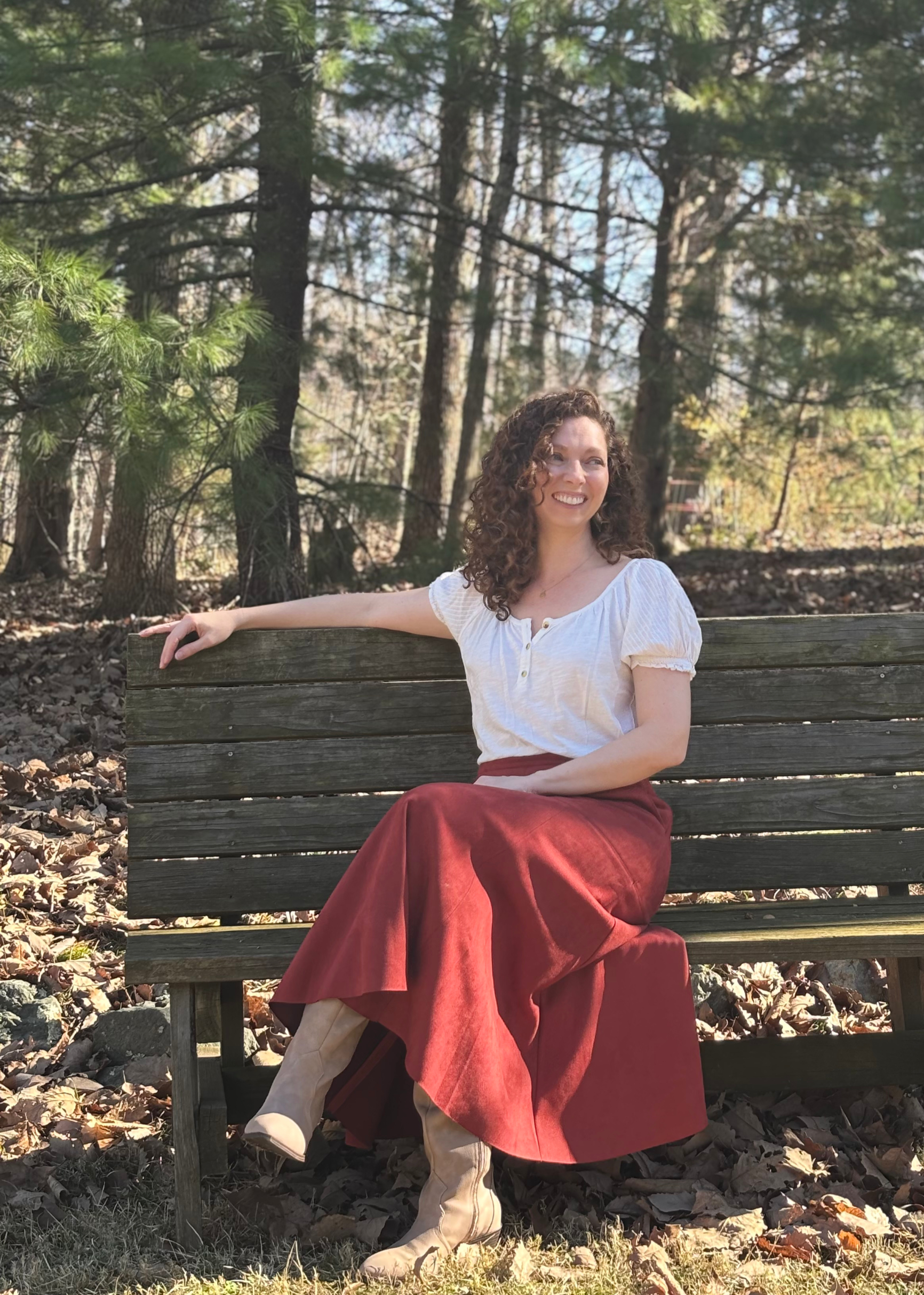 Raven Bonniwell seated on a wooden bench in a sunlit forest, wearing a white top and a red maxi skirt