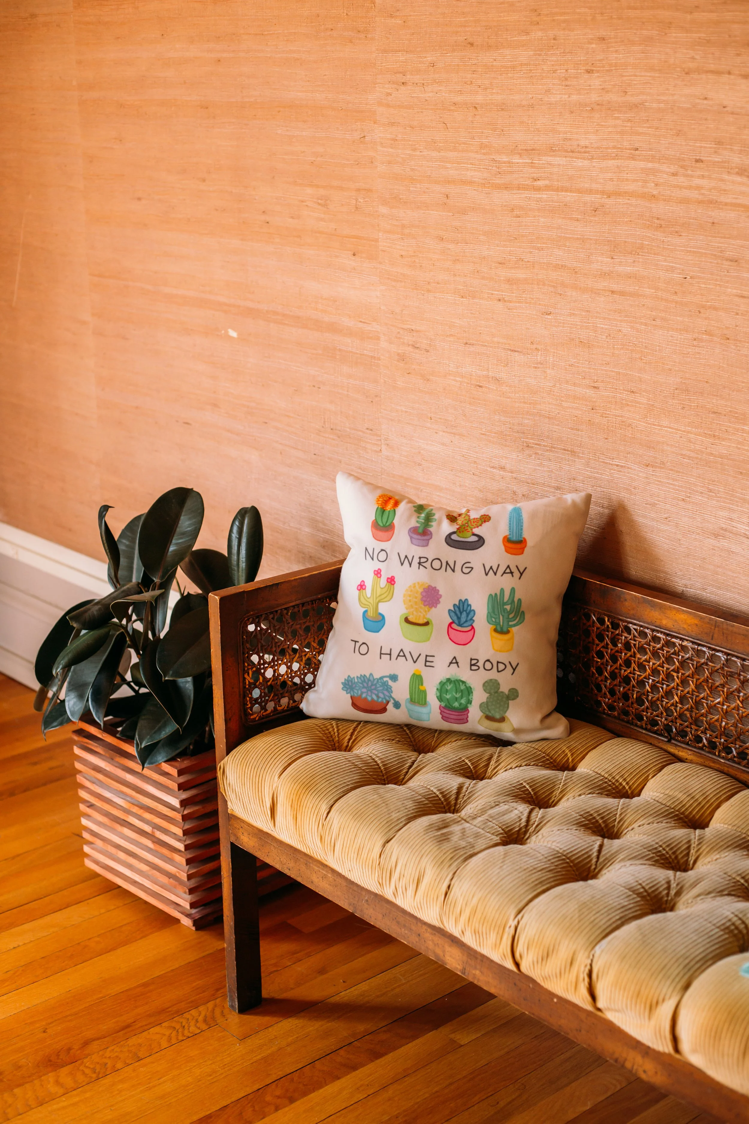 Cozy RealBeautifulYOU studio interior with a tufted bench, a "No Wrong Way to Have a Body" cactus-print pillow, and a rubber plant against a warm grasscloth wall