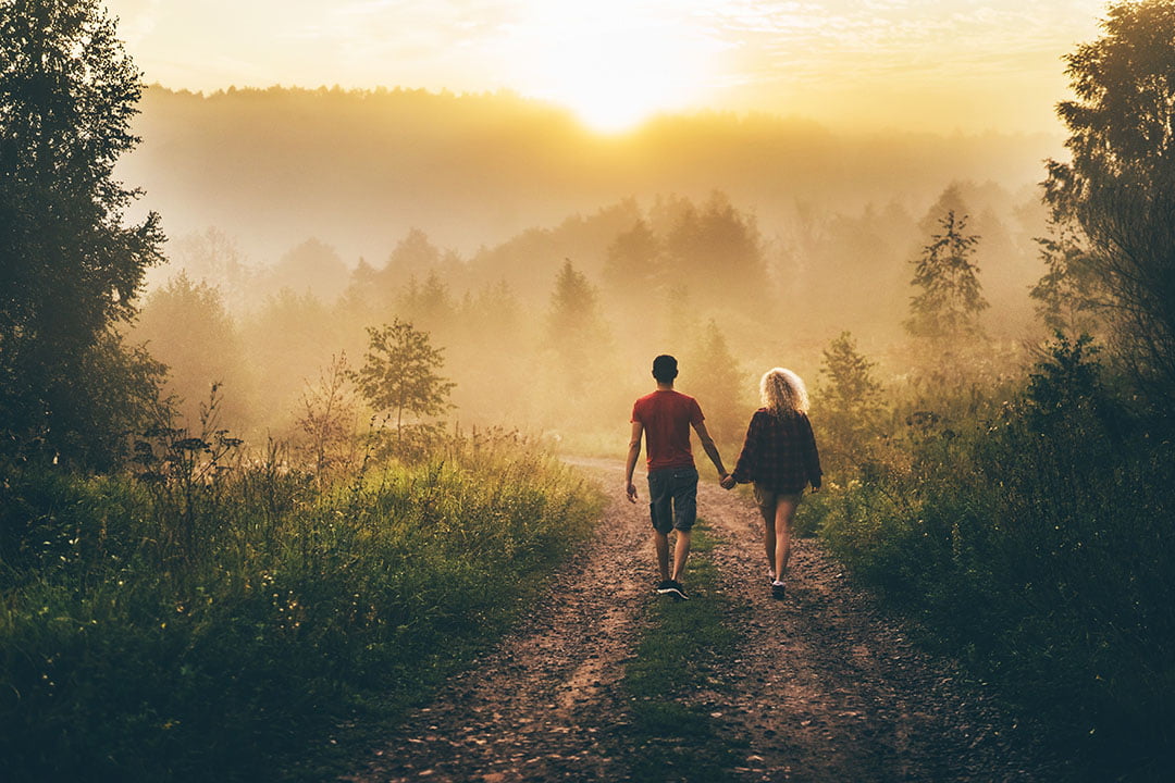 Couple holding hands walking down a misty forest path toward a golden sunset