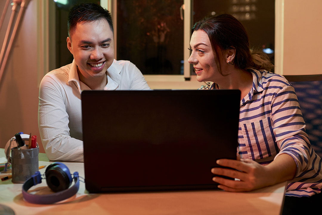 Man and woman smiling together while looking at a laptop screen in a warmly lit room