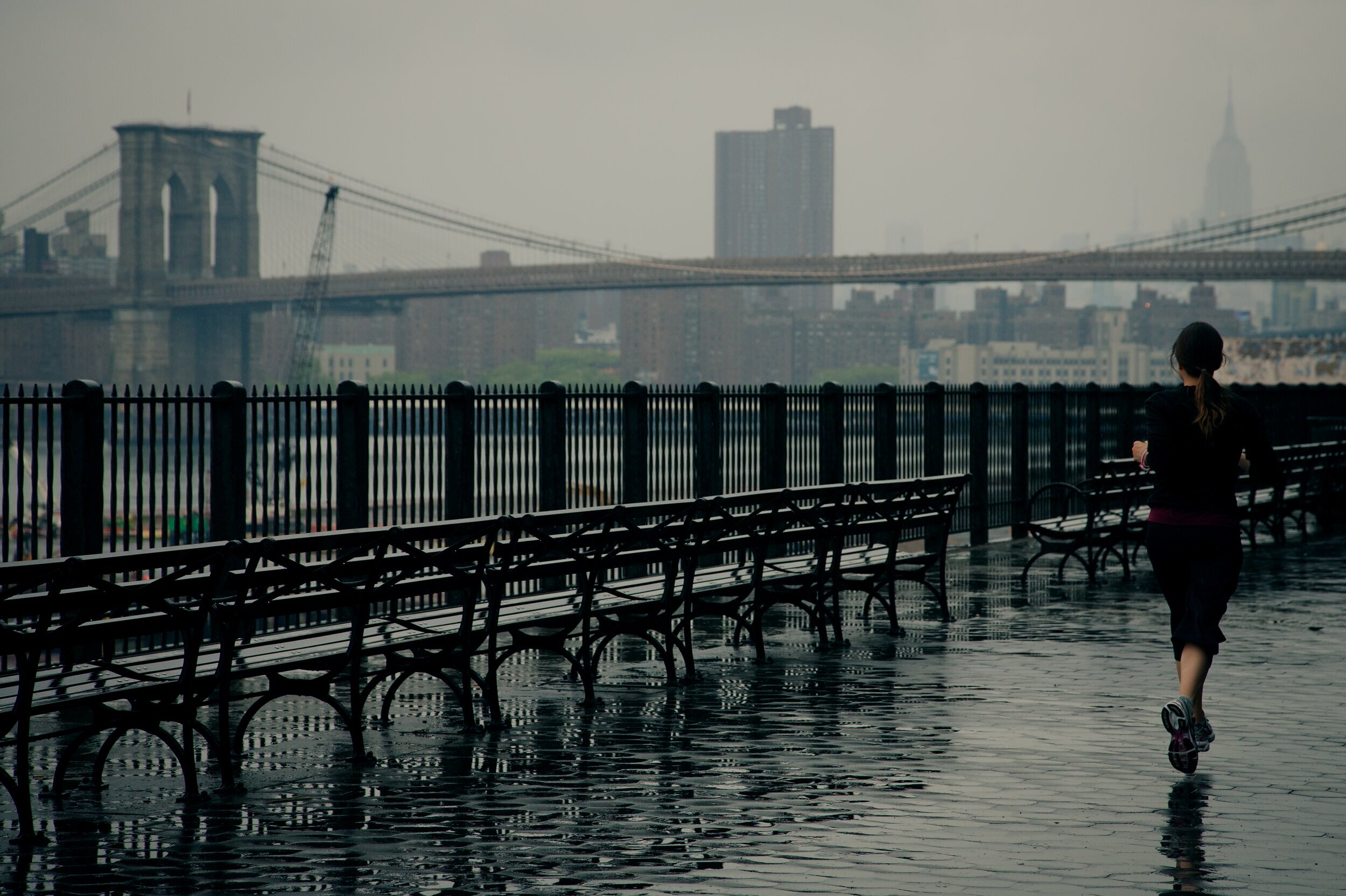 Runner on a wet promenade with the Brooklyn Bridge behind her