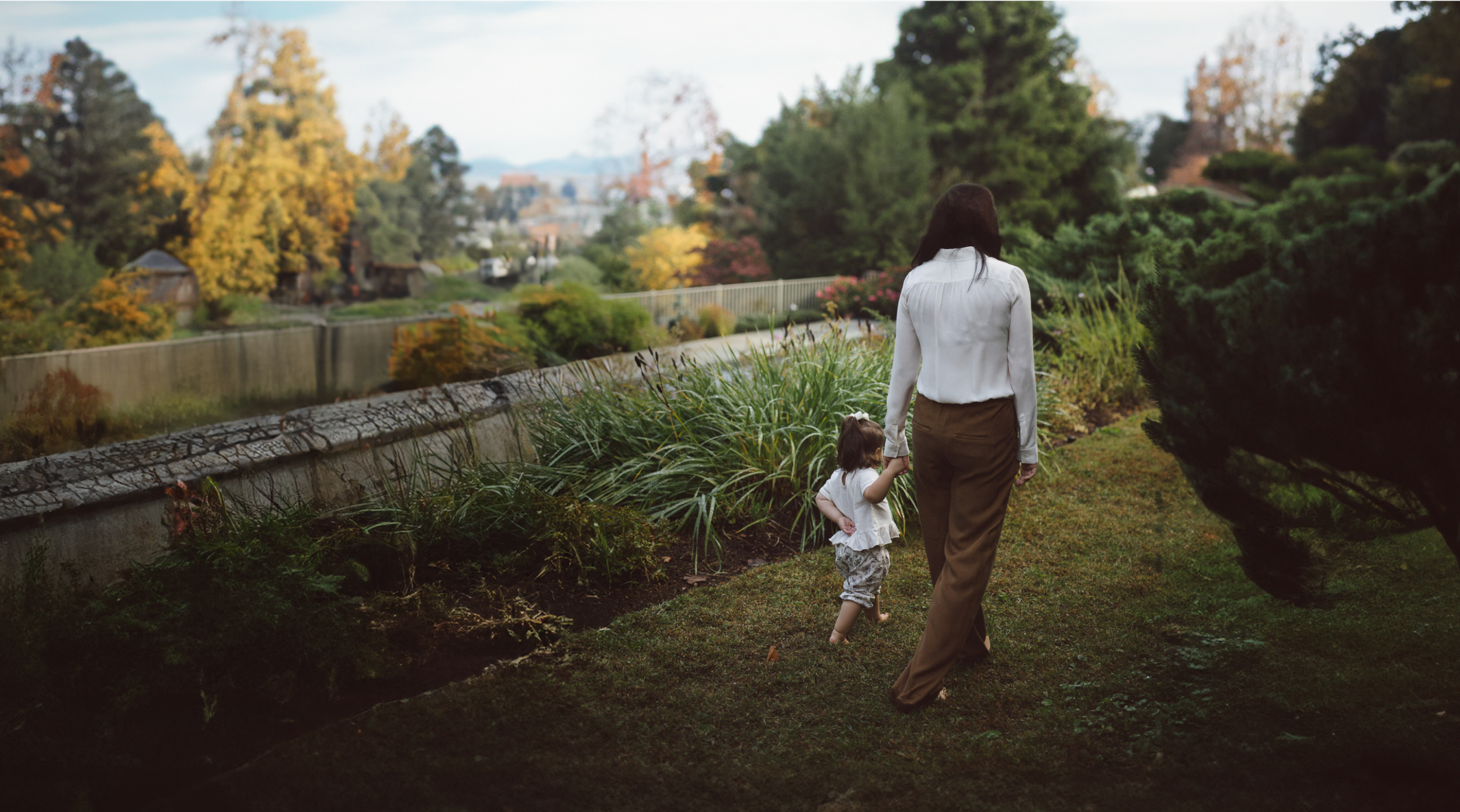 A mother and her young child walking together through a sunlit autumn garden