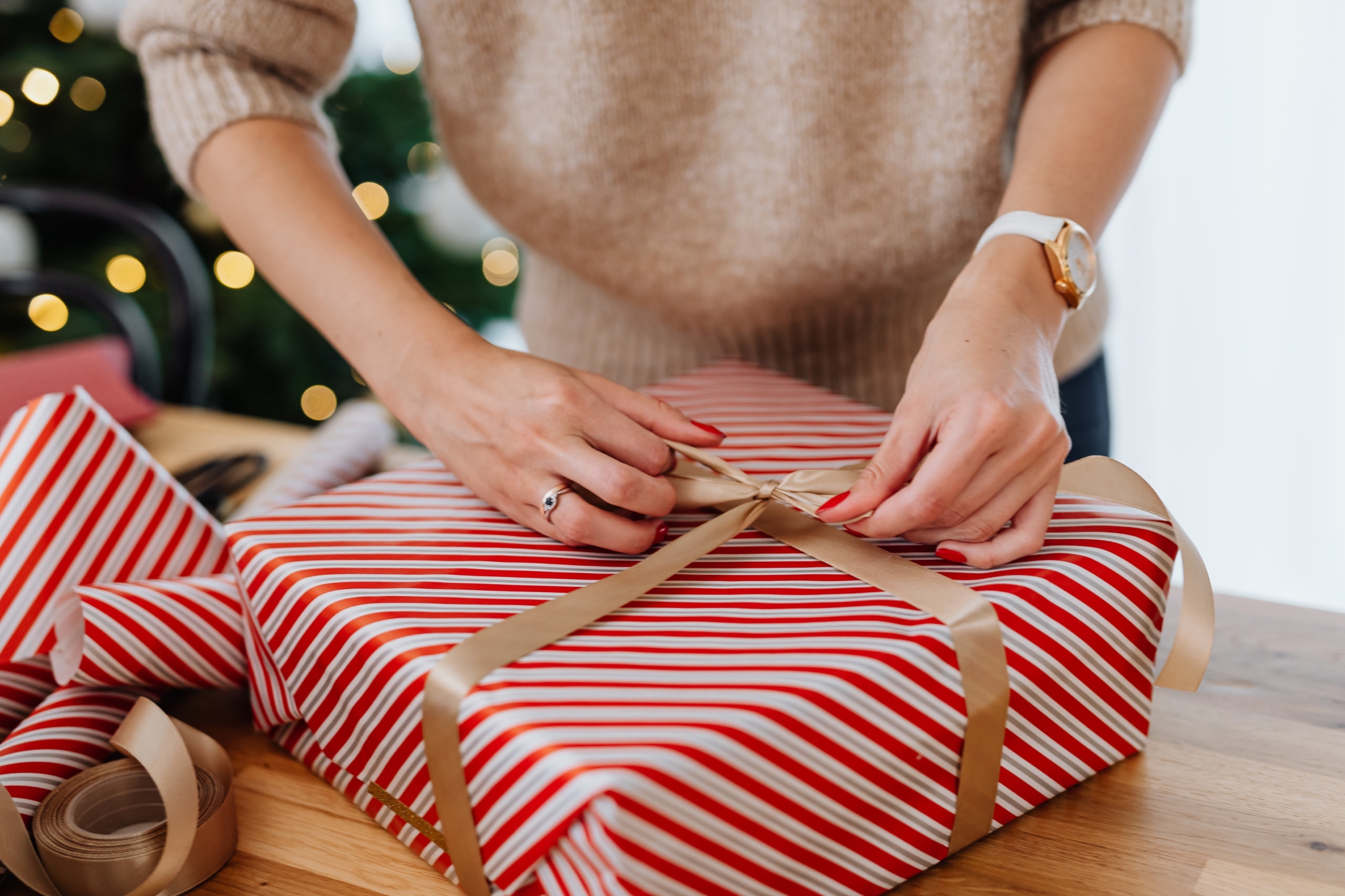 Hands wrapping a red and white striped gift with gold ribbon beside a lit holiday tree