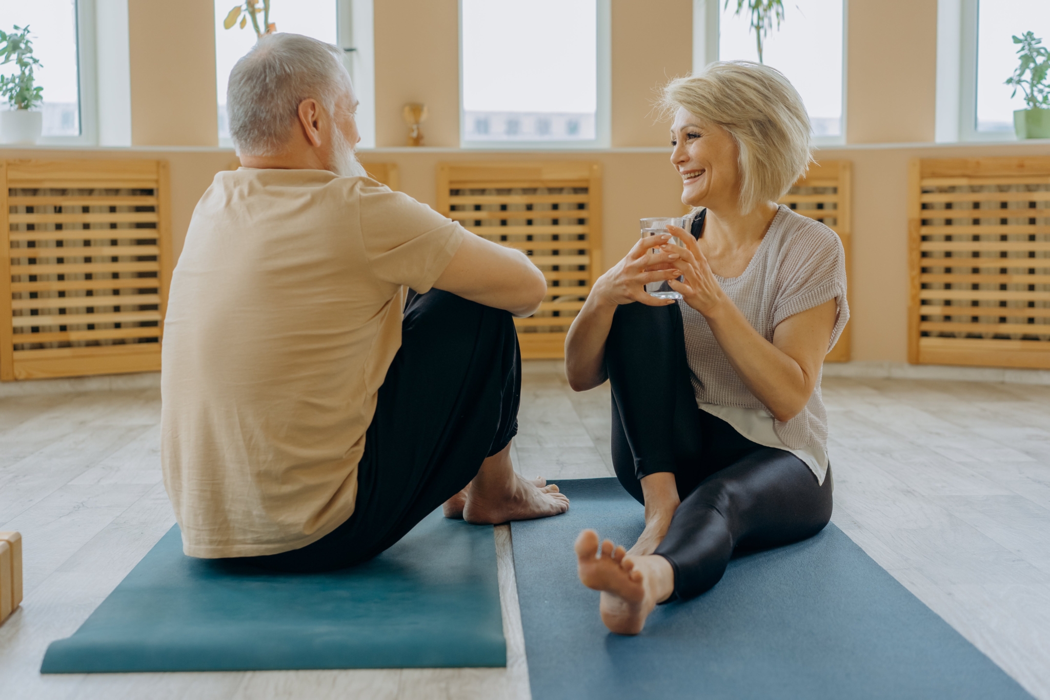 An older man and woman sitting on yoga mats in a bright studio, chatting and smiling
