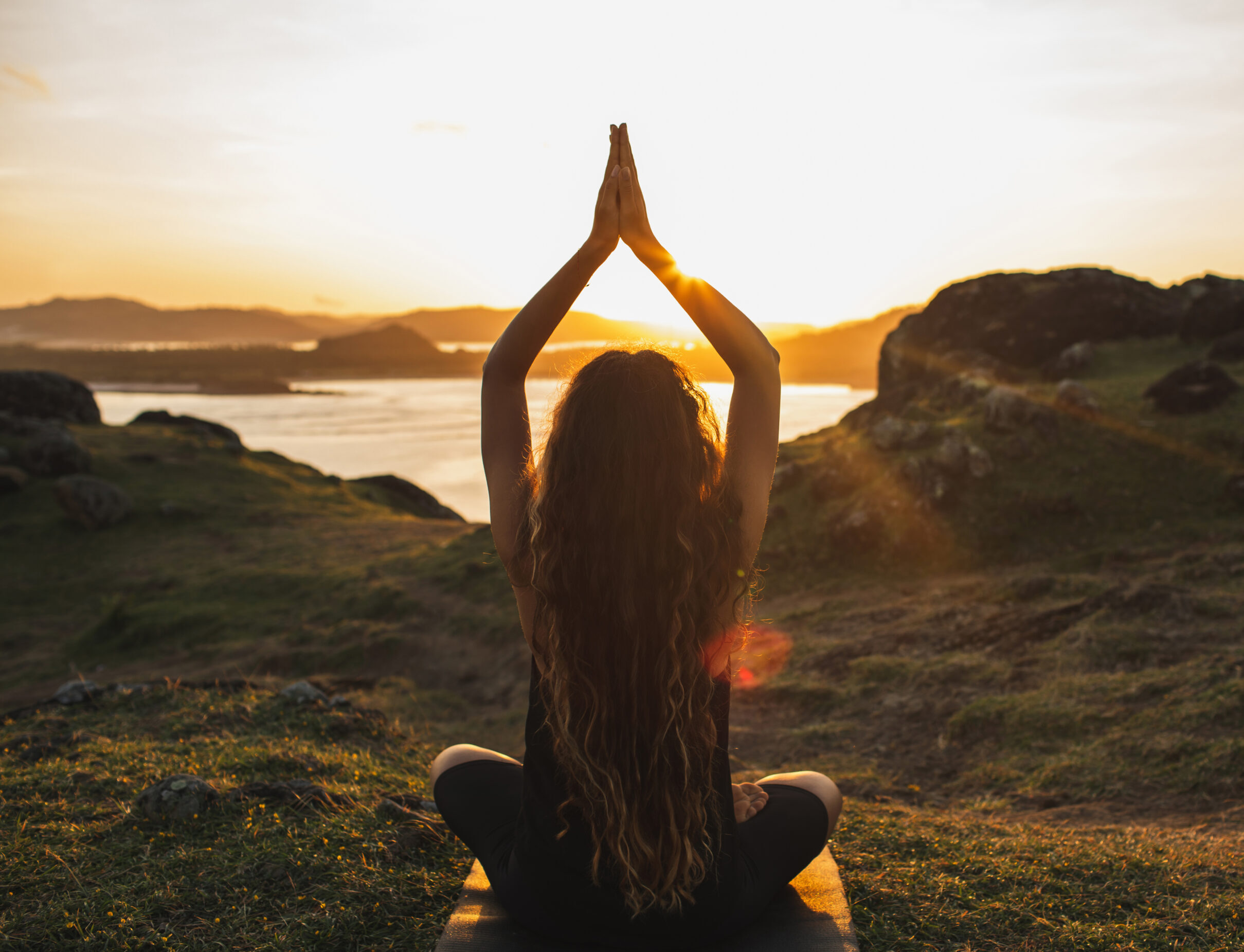 A woman seated cross-legged with hands in namaste overhead, silhouetted against a golden sunrise over mountains and water