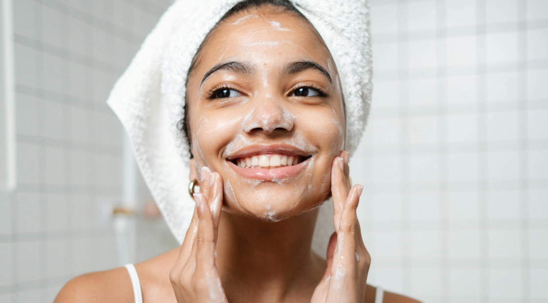 A smiling woman with a towel wrapped on her head washing her face with foamy cleanser