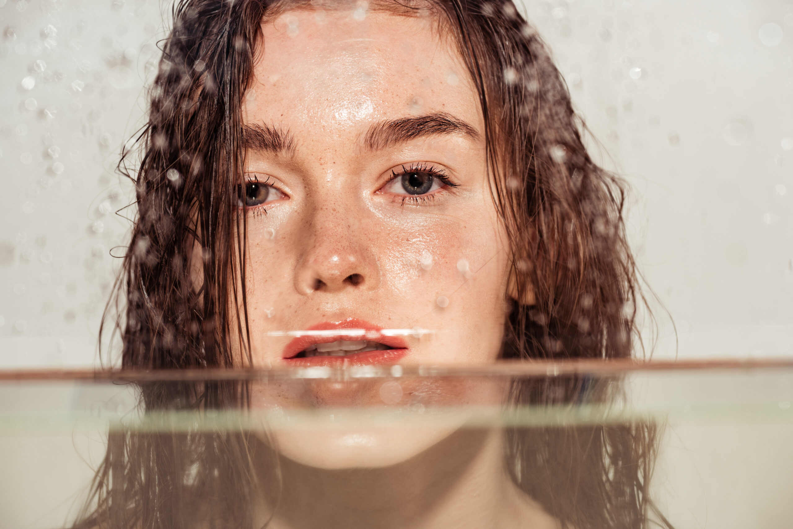 Close-up of a woman with wet hair and dewy skin seen through water-dropletted glass