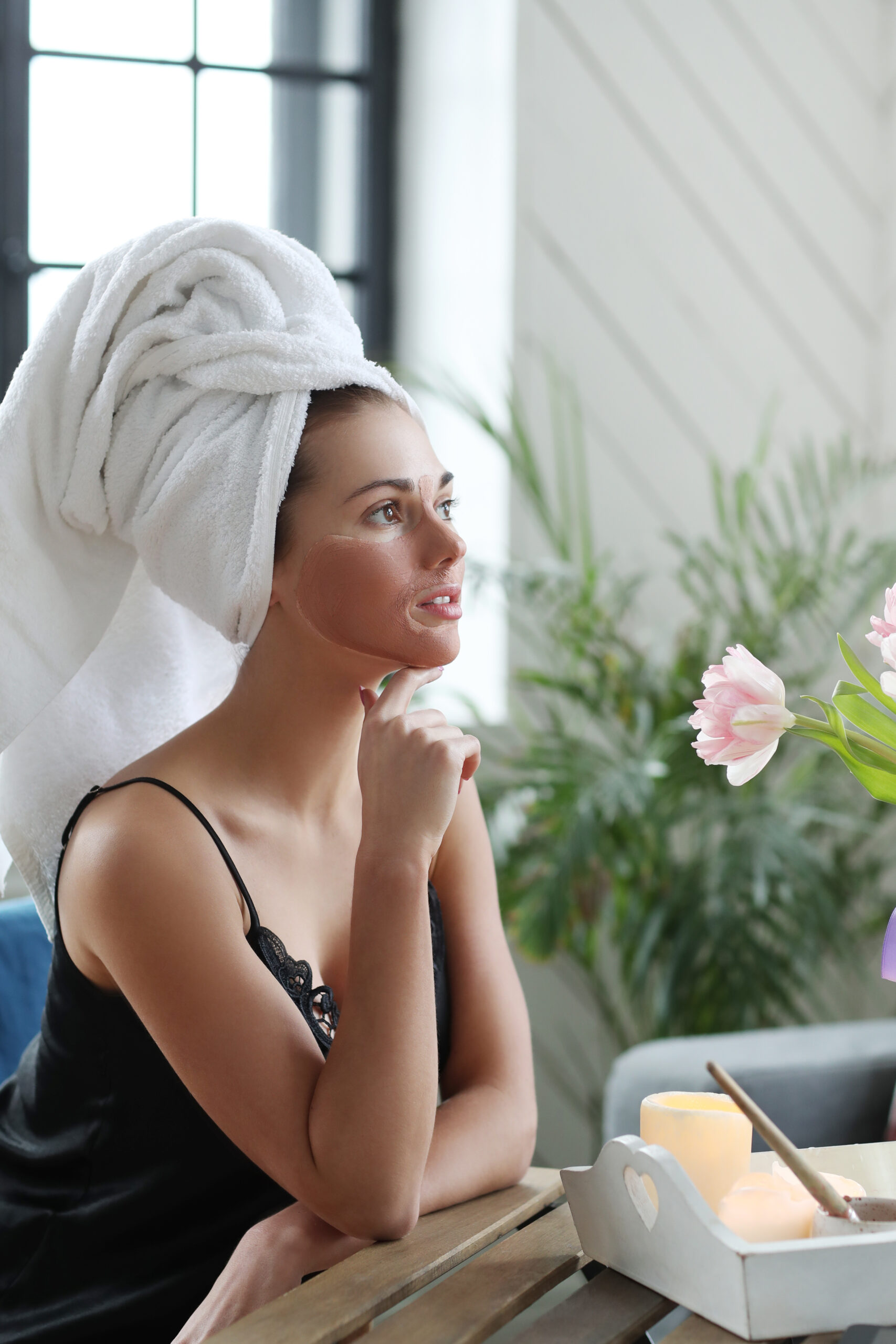 Woman with a towel on her head and under-eye patches seated by a window with flowers and candles