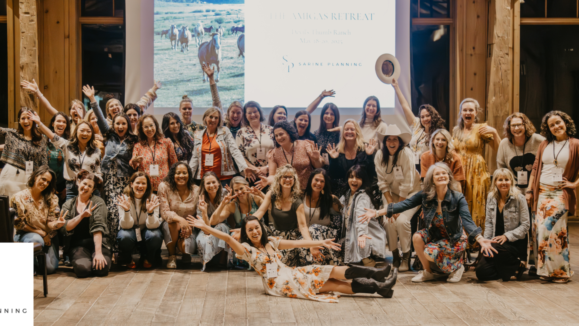 A large group of women posing enthusiastically at The Amigas Retreat, with Sarine Planning branding projected behind them