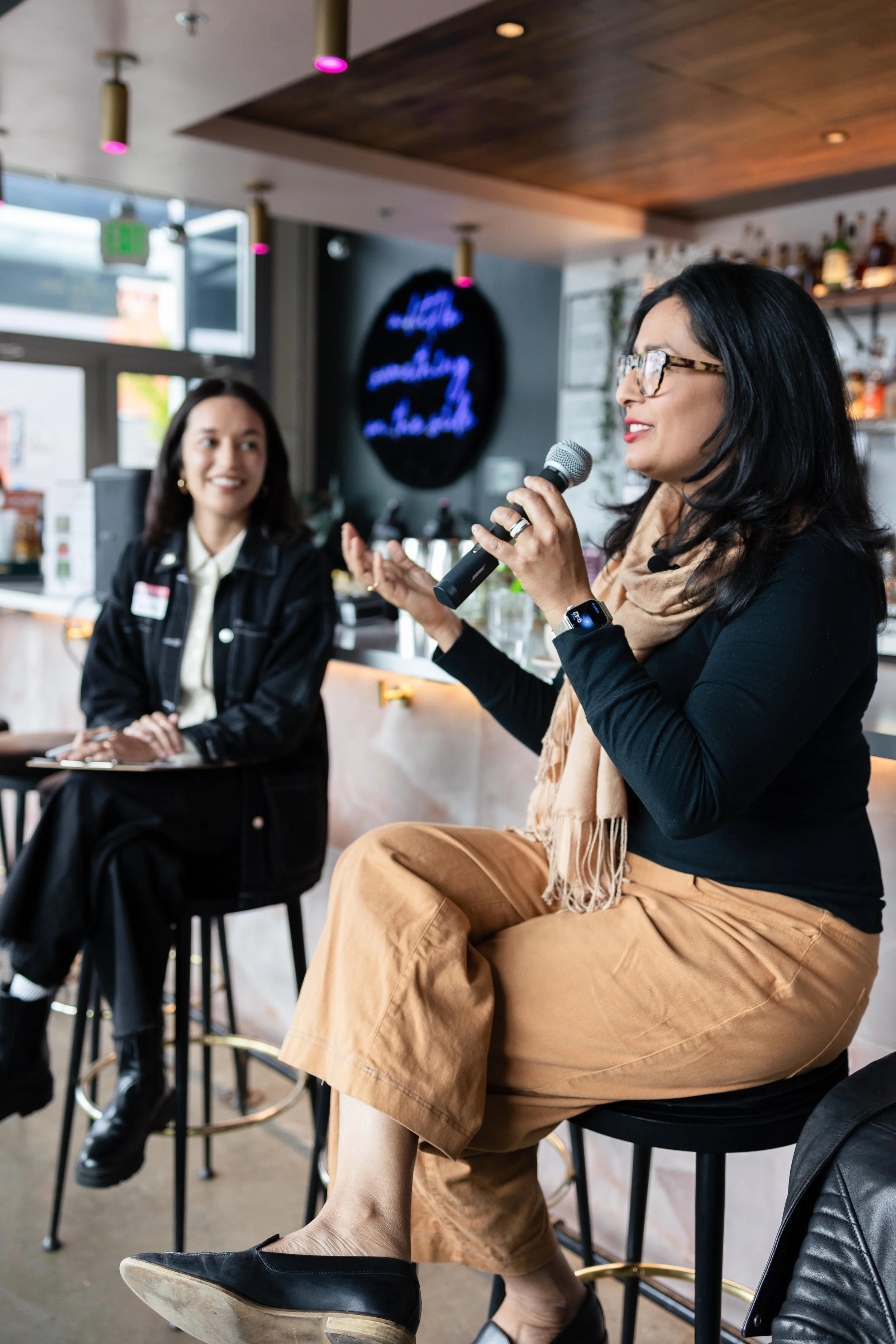 A woman with glasses and a tan scarf speaking into a microphone at a bar venue, another woman seated beside her