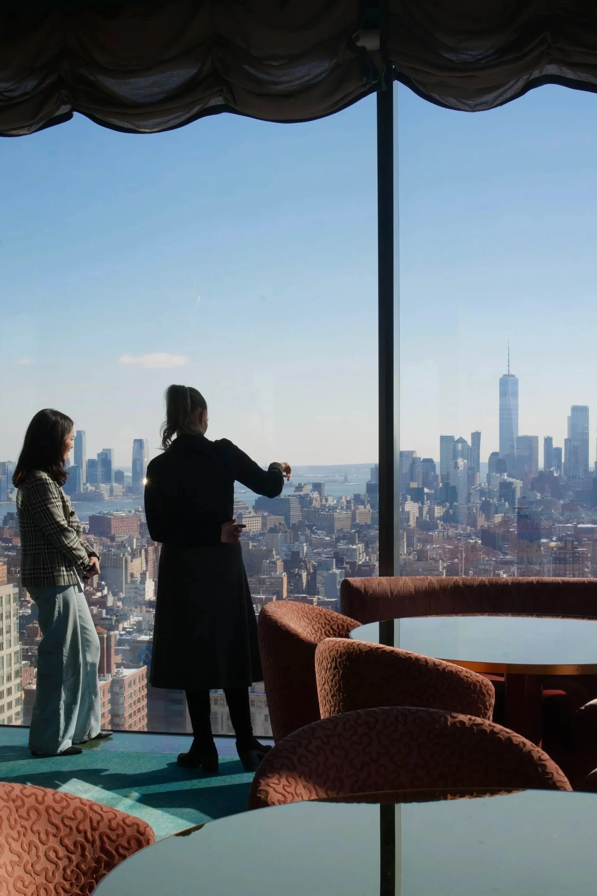 Two women silhouetted against floor-to-ceiling windows overlooking the lower Manhattan skyline in an upscale lounge