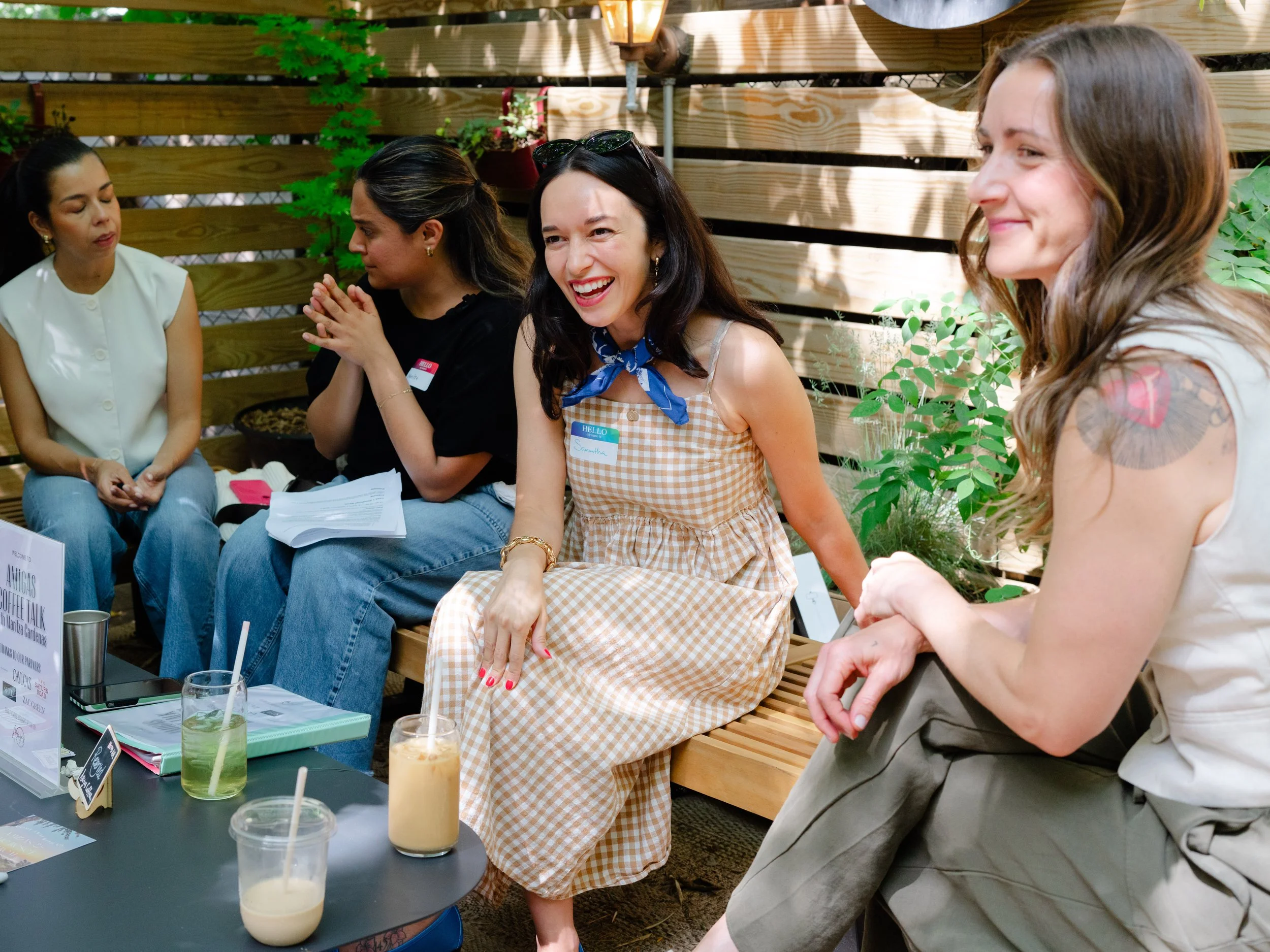 A group of women laughing at a casual outdoor event, wearing name tags and holding iced drinks