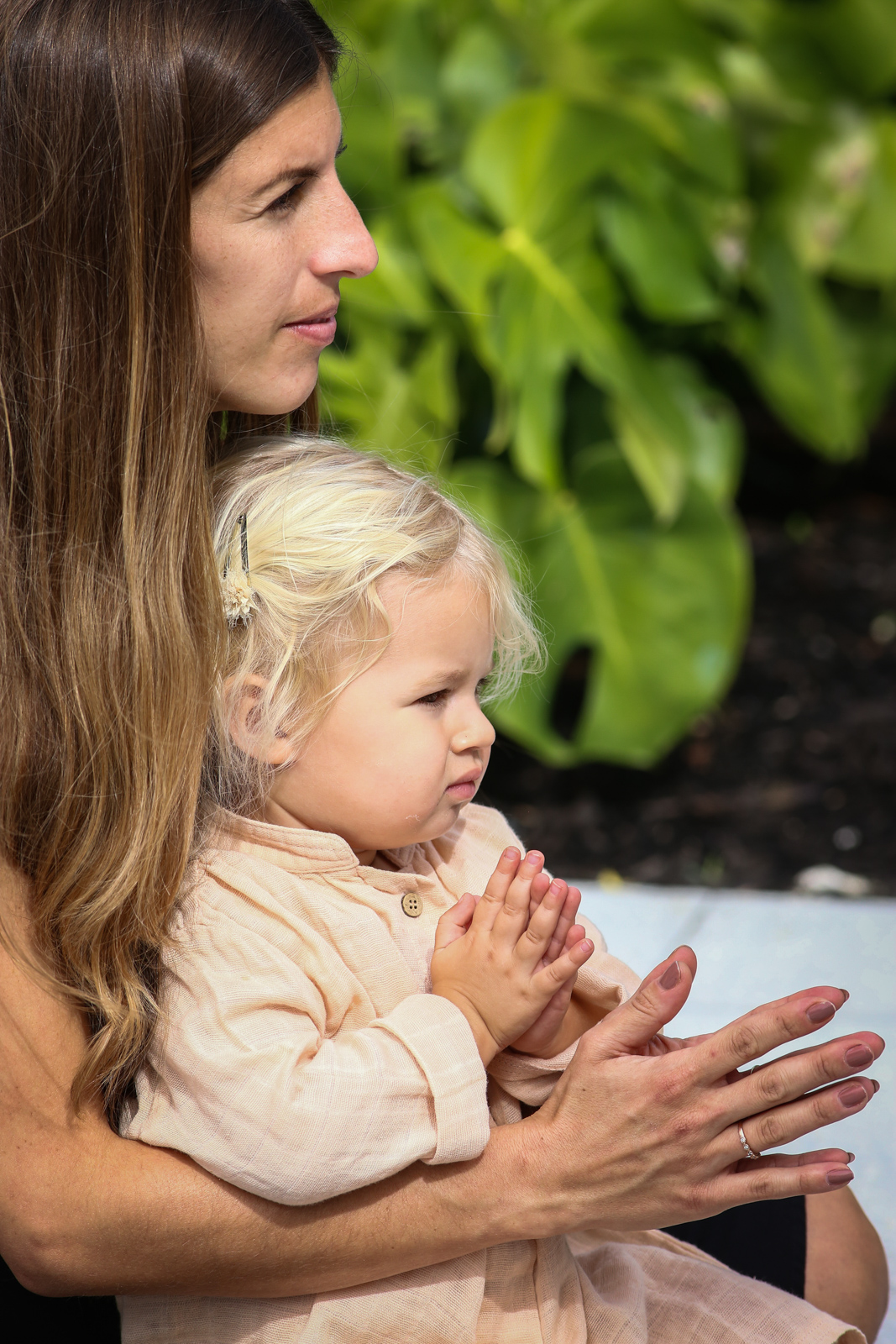 Mother holding a toddler on her lap at an outdoor yoga session, both clapping