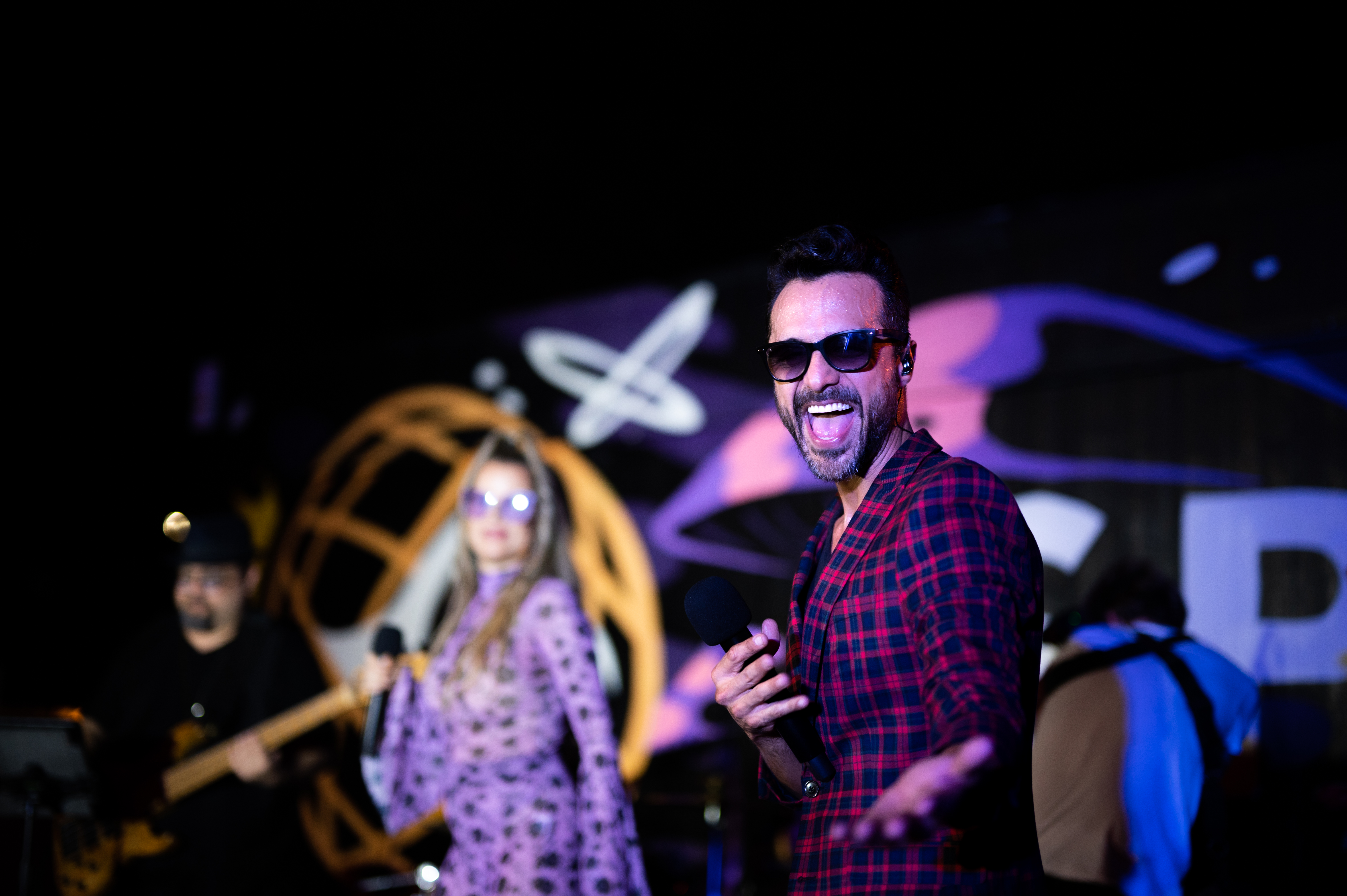 Performer in plaid shirt and sunglasses singing on stage at night at a SEED event