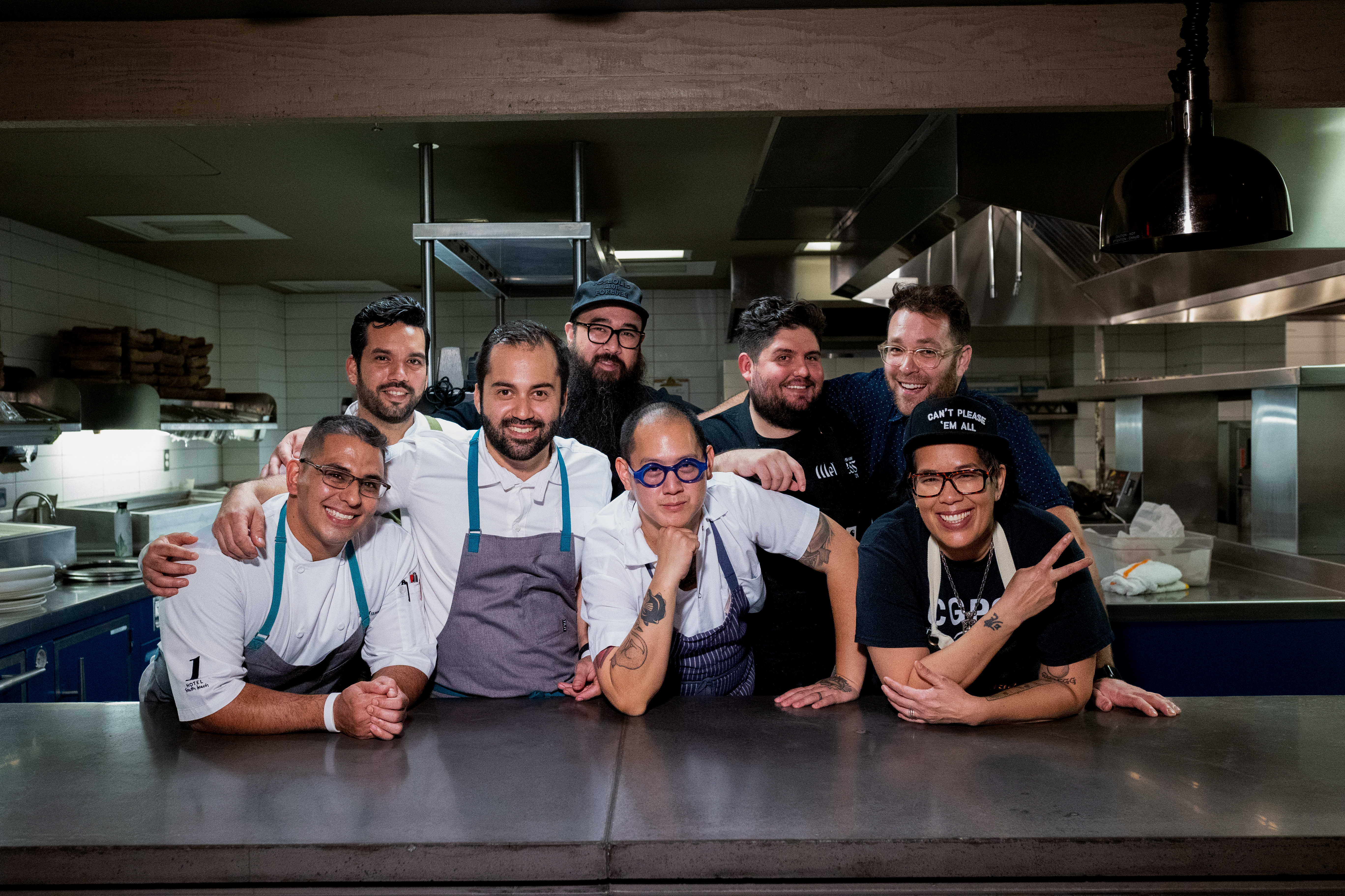 Eight chefs posing behind a kitchen prep counter