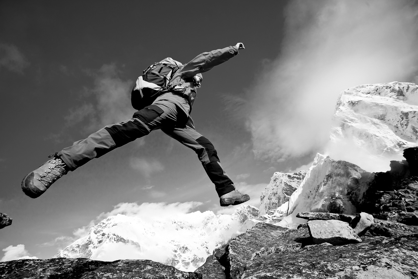 A hiker leaping between rocks on a mountain summit, snowy peaks behind — black and white