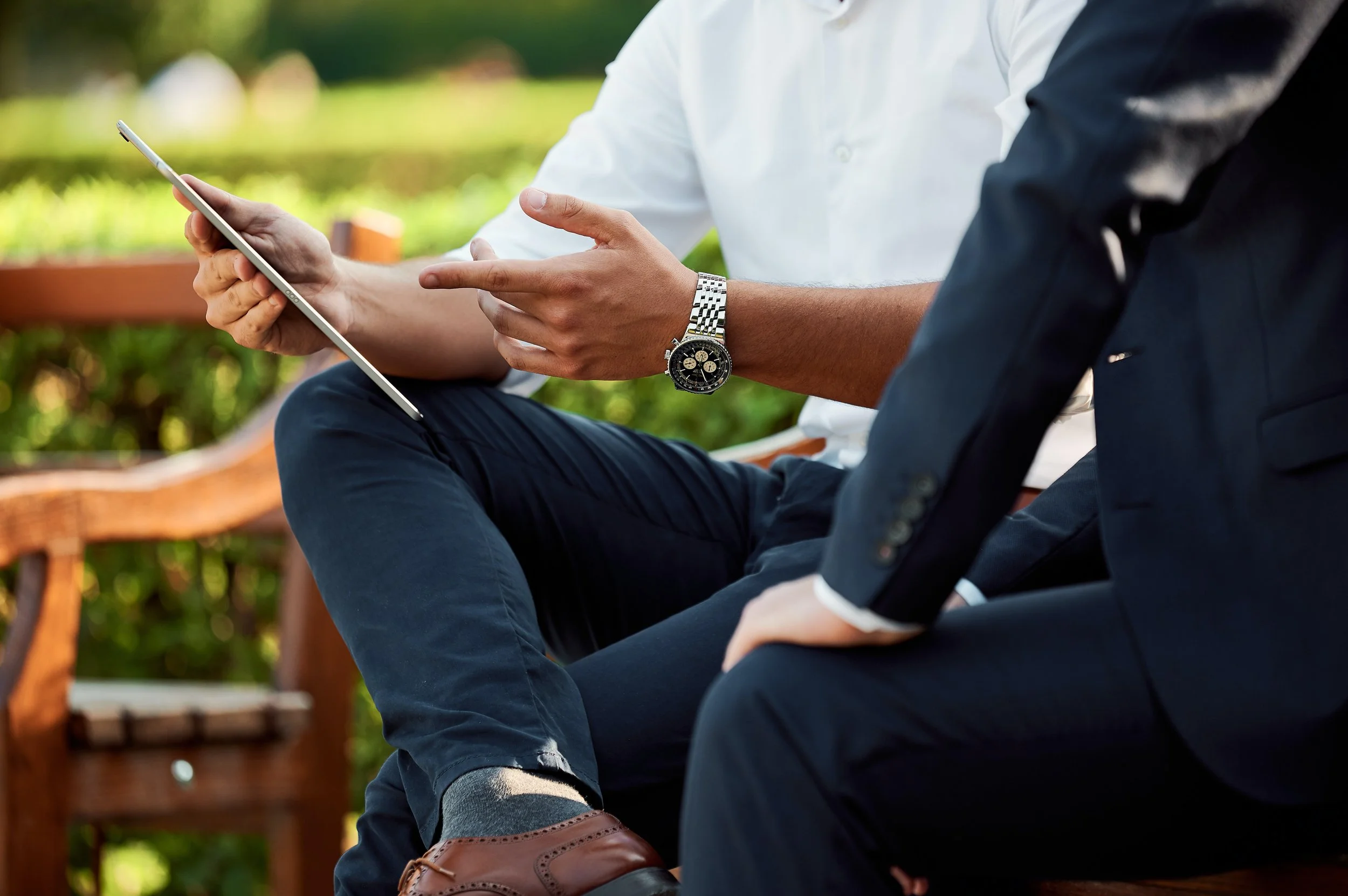 Two people sitting on a wooden bench, one holding a tablet, wearing a white shirt and watch, the other in a dark suit, outdoors with greenery