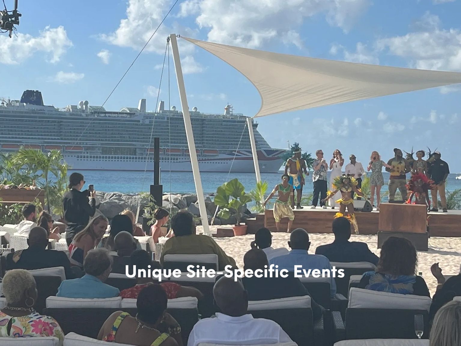 Outdoor beachside event with performers on stage, seated audience, and a cruise ship in the background