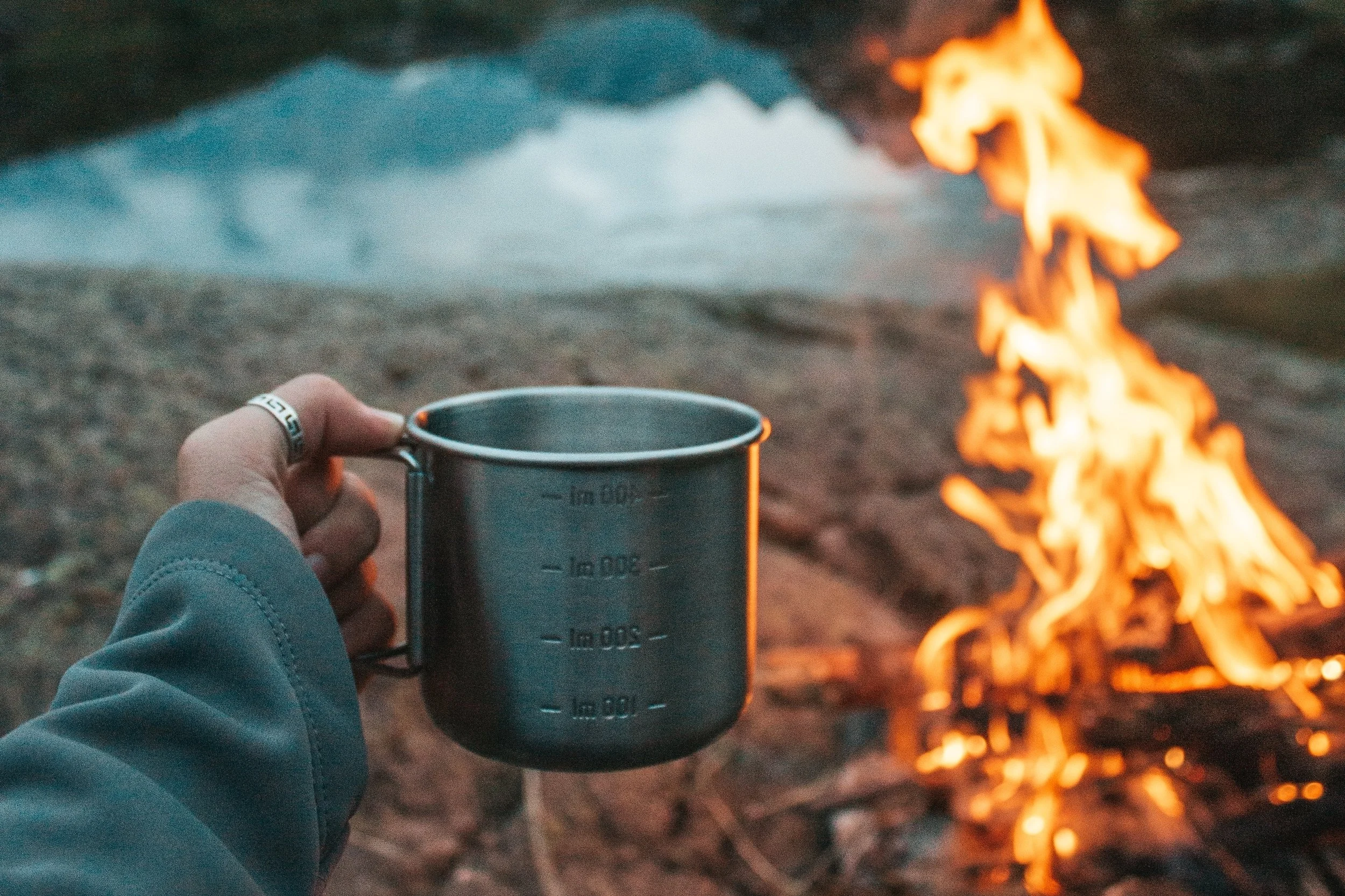 Hand holding a metal camping mug in front of a campfire outdoors