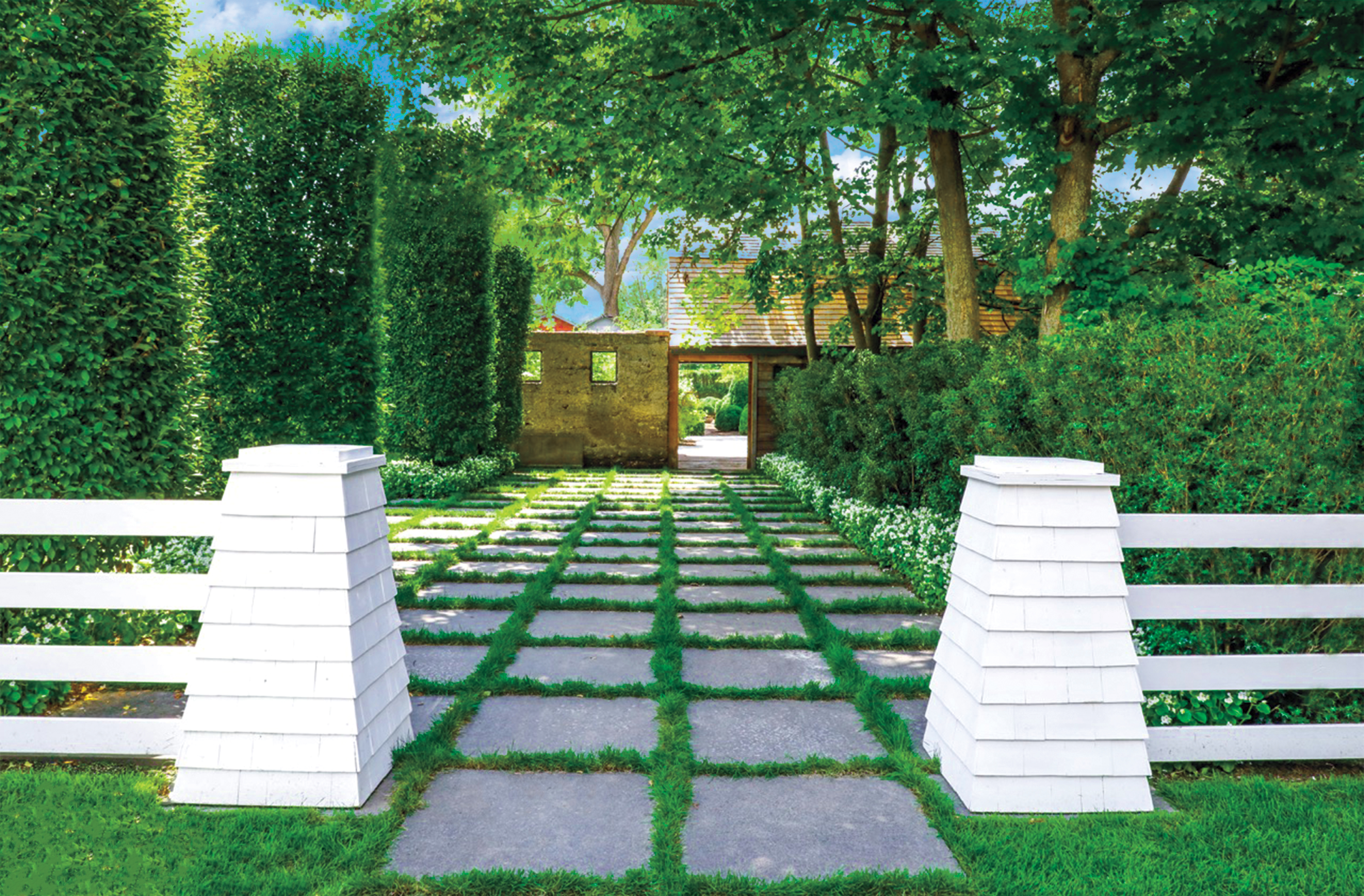 Lush garden pathway with white fence posts, stone pavers set in grass, tall hedges and a gate leading to a cottage