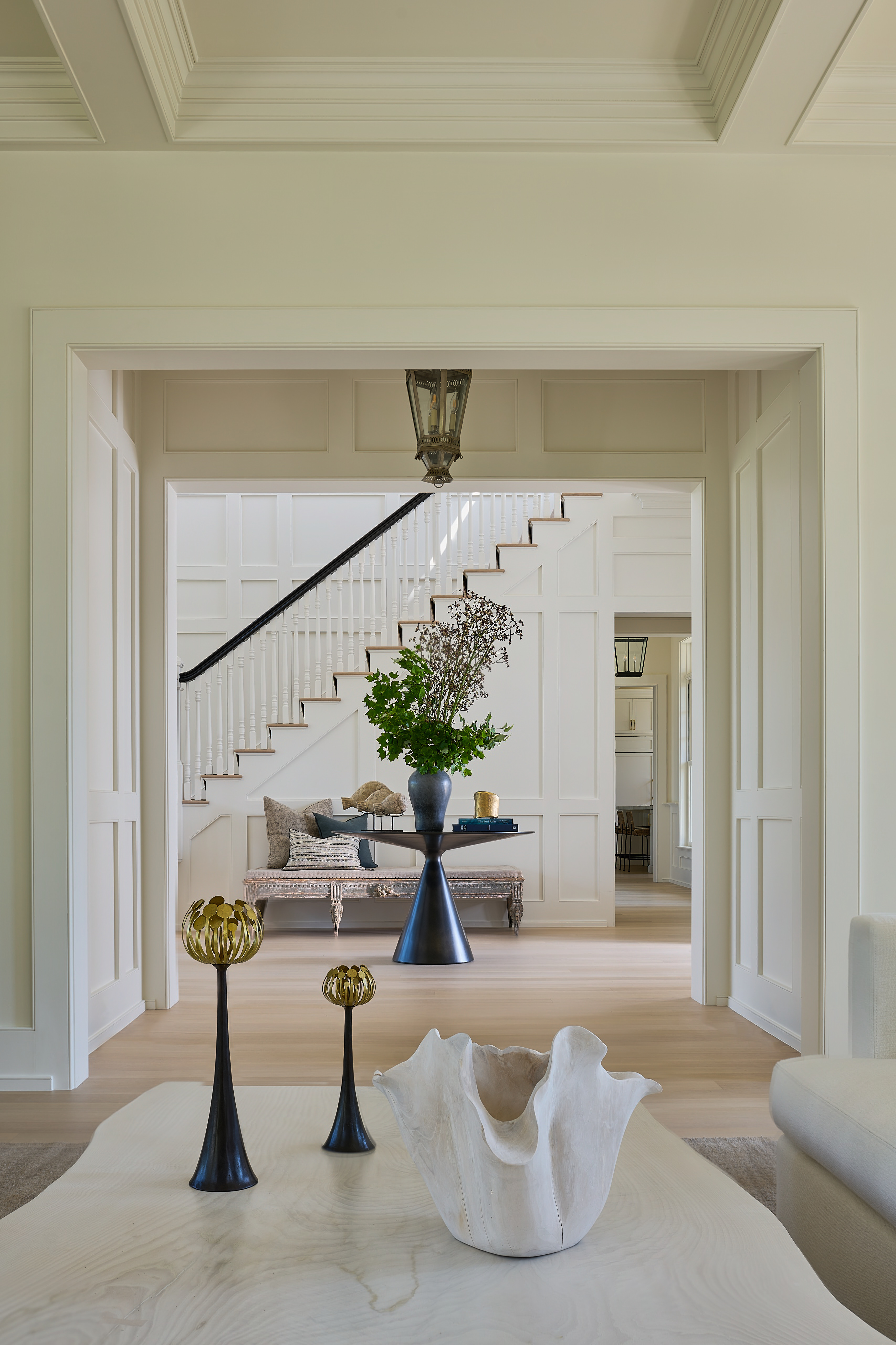 Elegant home foyer viewed through an archway with staircase, black pedestal table with floral arrangement and coffered ceiling