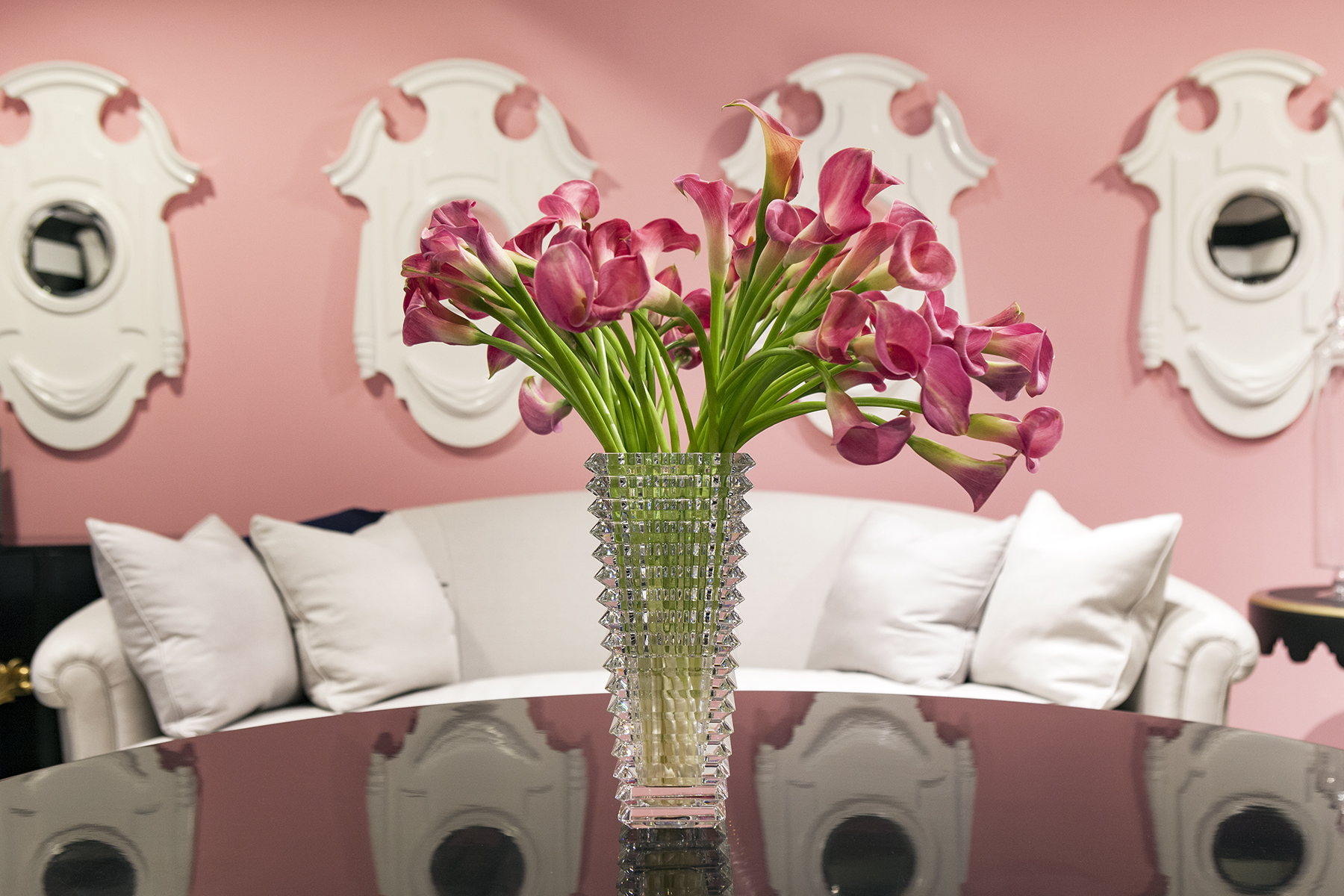 Pink-walled interior vignette: crystal vase of pink calla lilies on a reflective black table, white curved sofa and ornate white wall mirrors