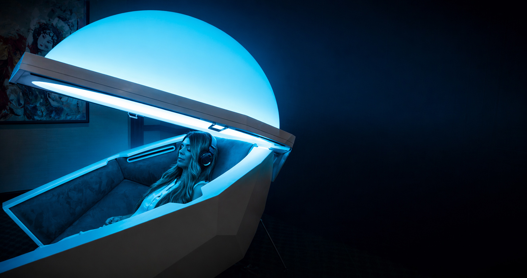 A woman with headphones reclining inside an open Somadome wellness pod, illuminated by blue LED light.