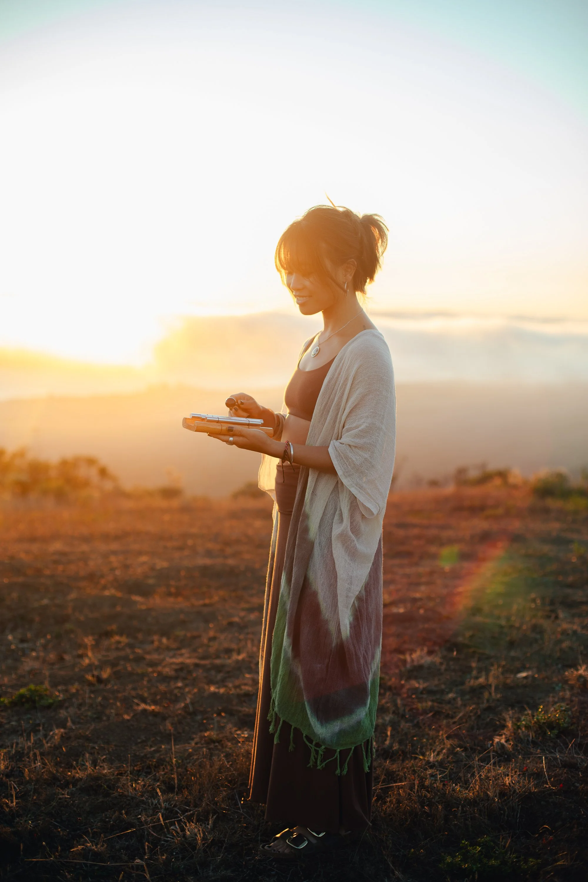Erica Nguyễn on a hillside at sunset holding a sound instrument, wearing a flowing kimono wrap